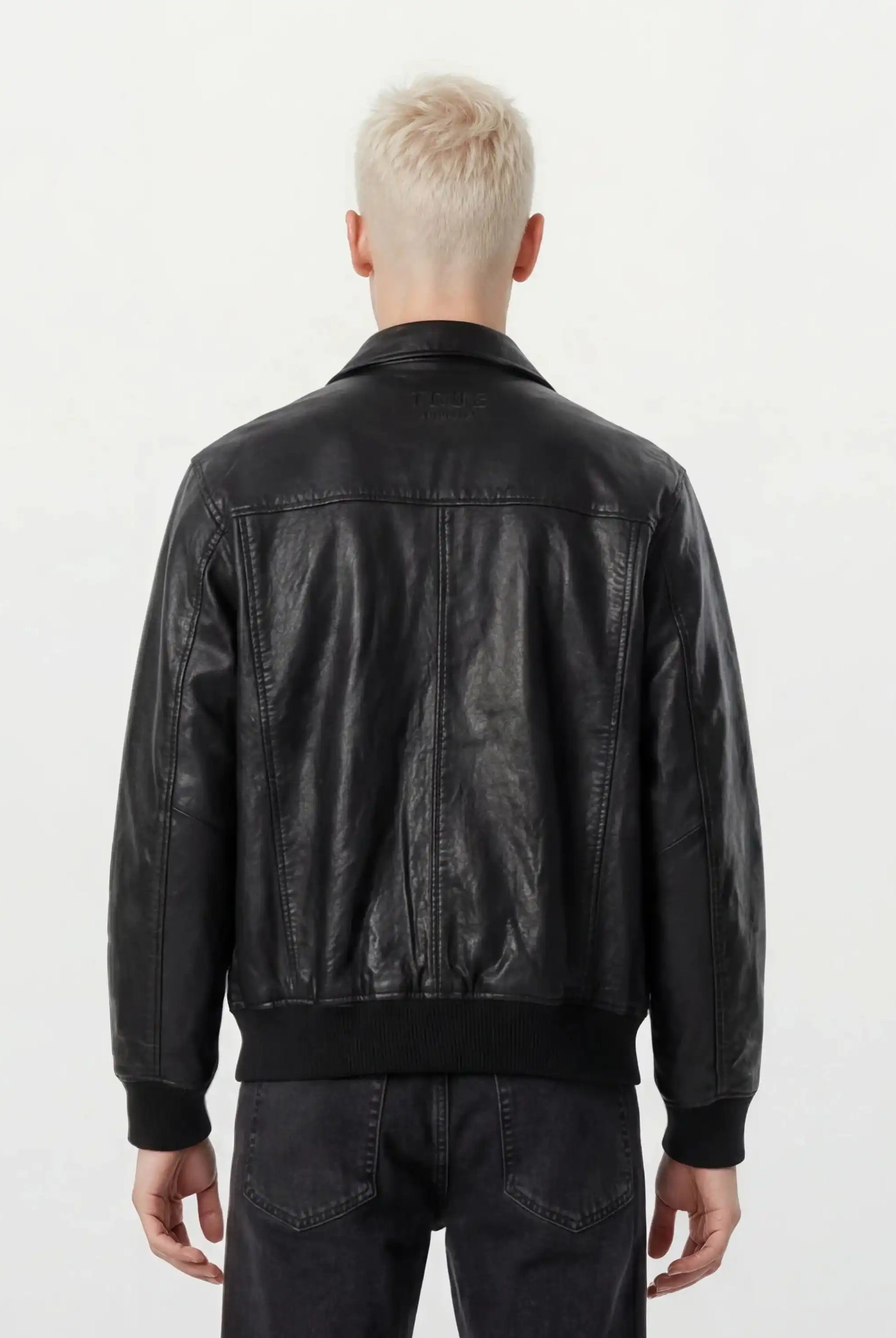 Eine Person mit kurzen, platinblonden Haaren ist von hinten zu sehen und trägt eine trueprodigy Oversize-Lederjacke mit Stehkragen, 3D-Silikonlogo & Retro-Details und dunkle Jeans vor einem schlichten, hellen Hintergrund.