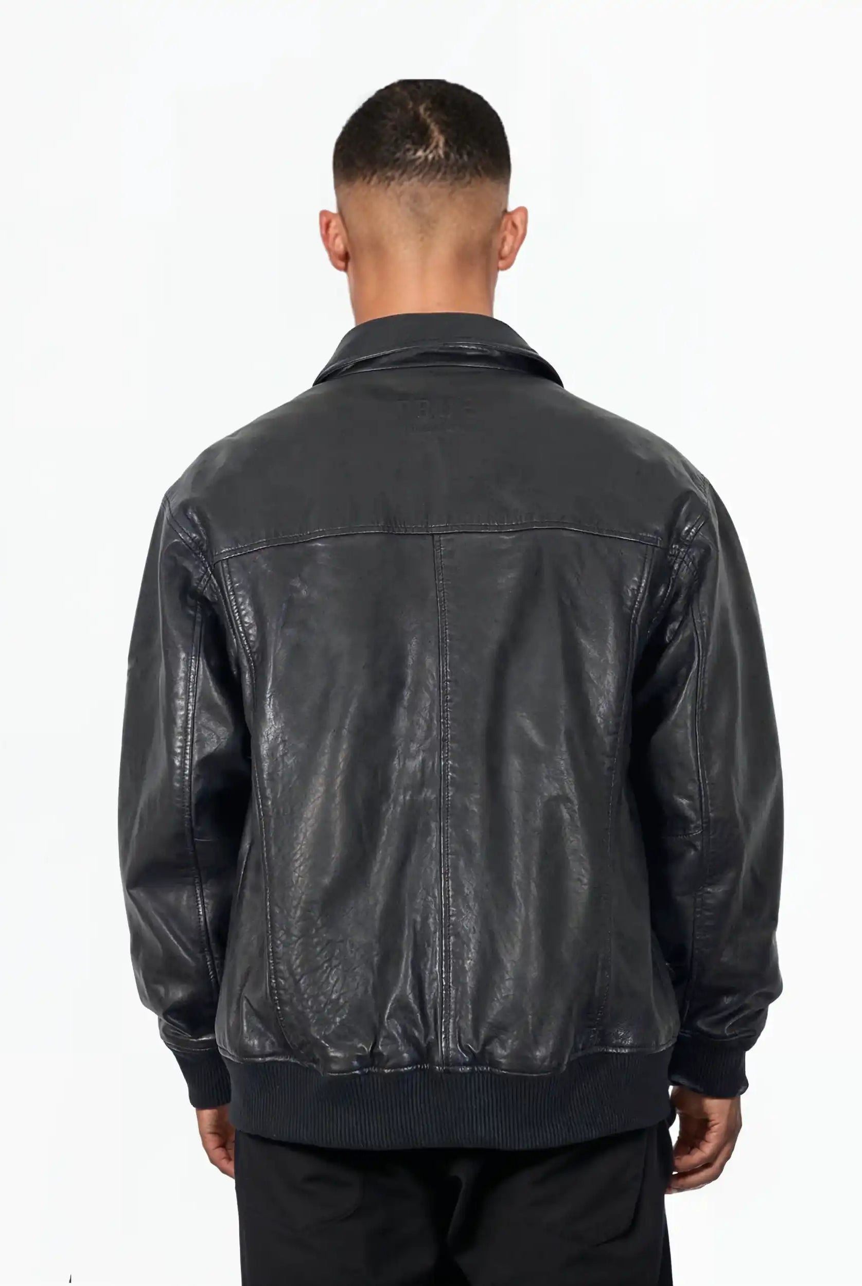 Eine Person steht mit dem Rücken zur Kamera und trägt die trueprodigy Ryder Oversize-Lederjacke mit Stehkragen, 3D-Silikonlogo & Retro-Details und schwarzer Hose vor einem schlichten weißen Hintergrund.