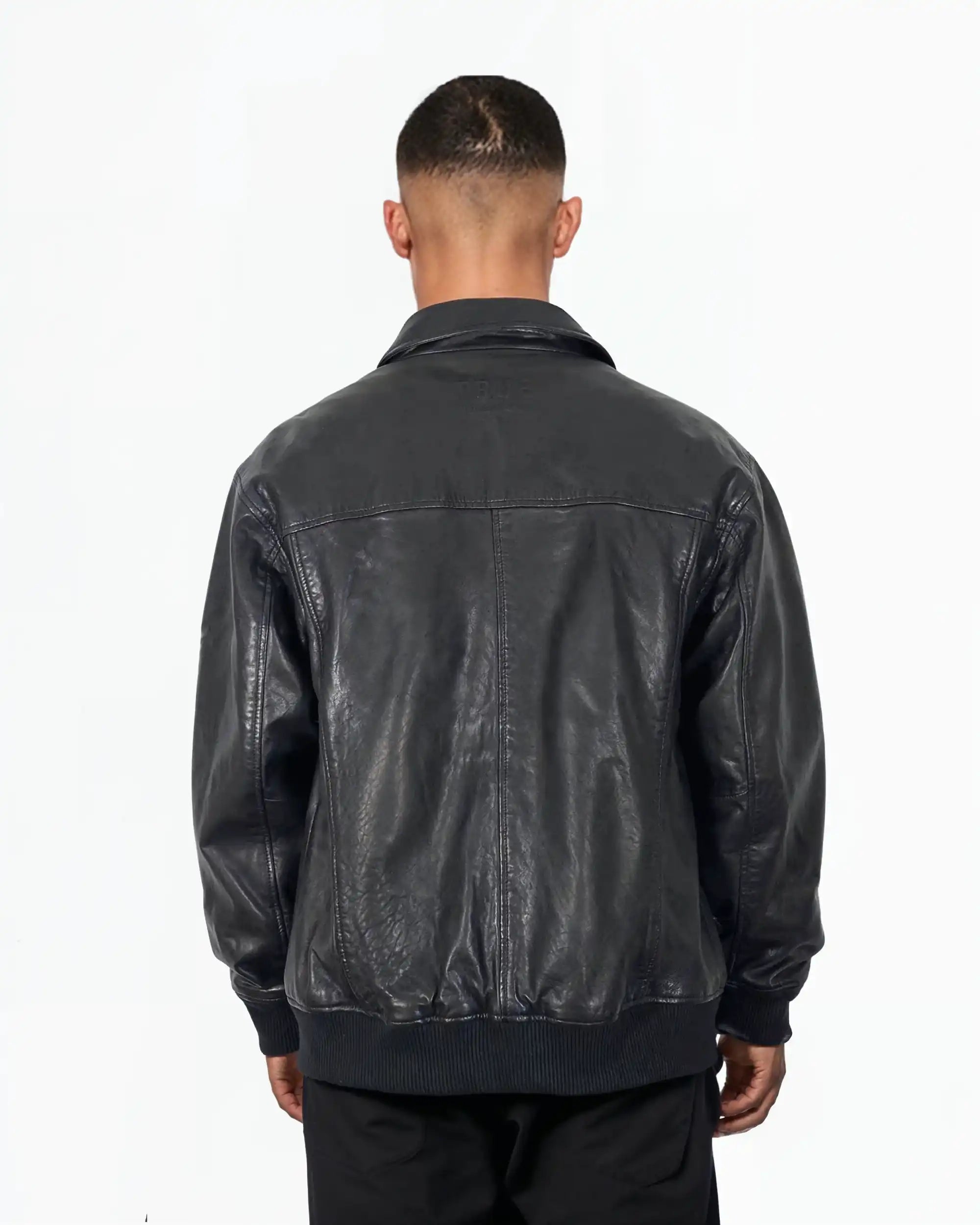 Eine Person steht mit dem Rücken zur Kamera und trägt die trueprodigy Ryder Oversize-Lederjacke mit Stehkragen, 3D-Silikonlogo & Retro-Details und schwarzer Hose vor einem schlichten weißen Hintergrund.