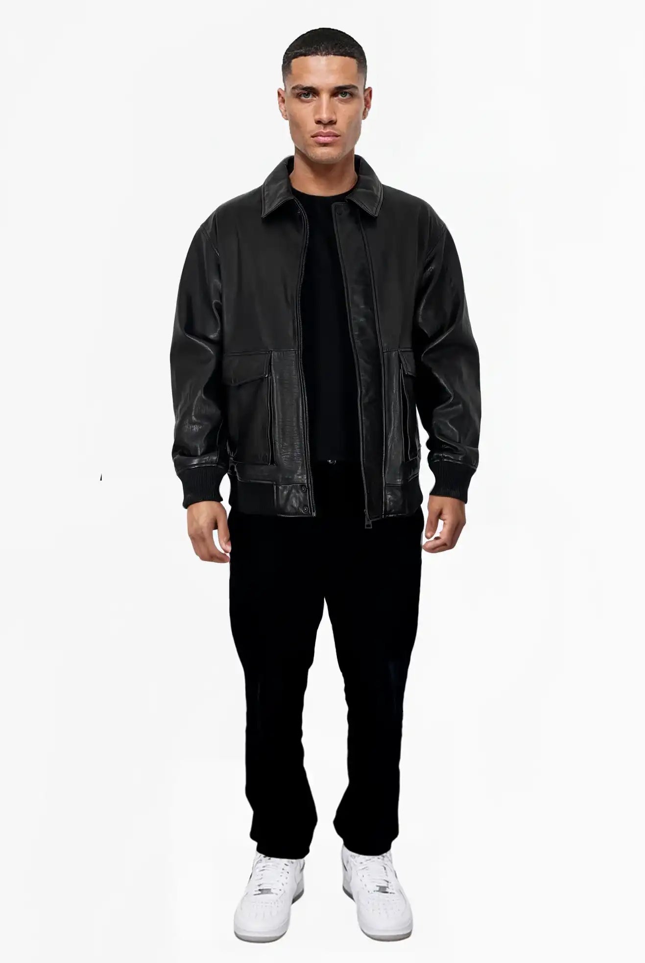 Ein Mann steht vor einem weißen Hintergrund und trägt die Ryder Oversize-Lederjacke mit Stehkragen, 3D-Silikonlogo & Retro-Details von trueprodigy, gepaart mit einer schwarzen Hose, einem schwarzen Hemd und weißen Turnschuhen. Seine Arme ruhen an den Seiten.