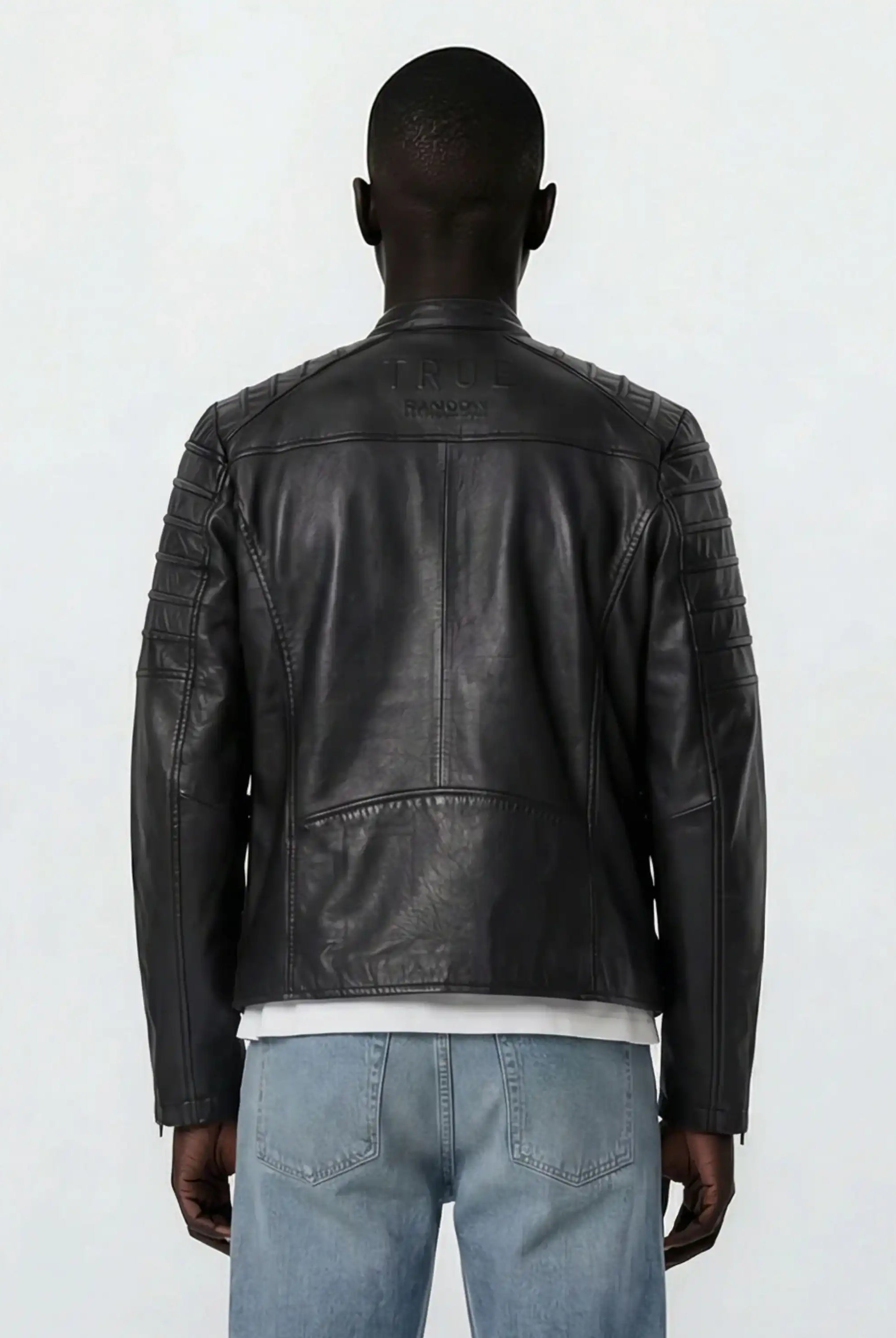 Eine Person steht mit dem Rücken zur Kamera und trägt eine schwarze trueprodigy Slim-Fit-Lederjacke mit Stehkragen & Zipper-Details mit abgesteppten Schultern und hellblauen Jeans. TRUE und TRANOLOGY sind in der Nähe des Kragens auf dem Rücken eingeprägt.