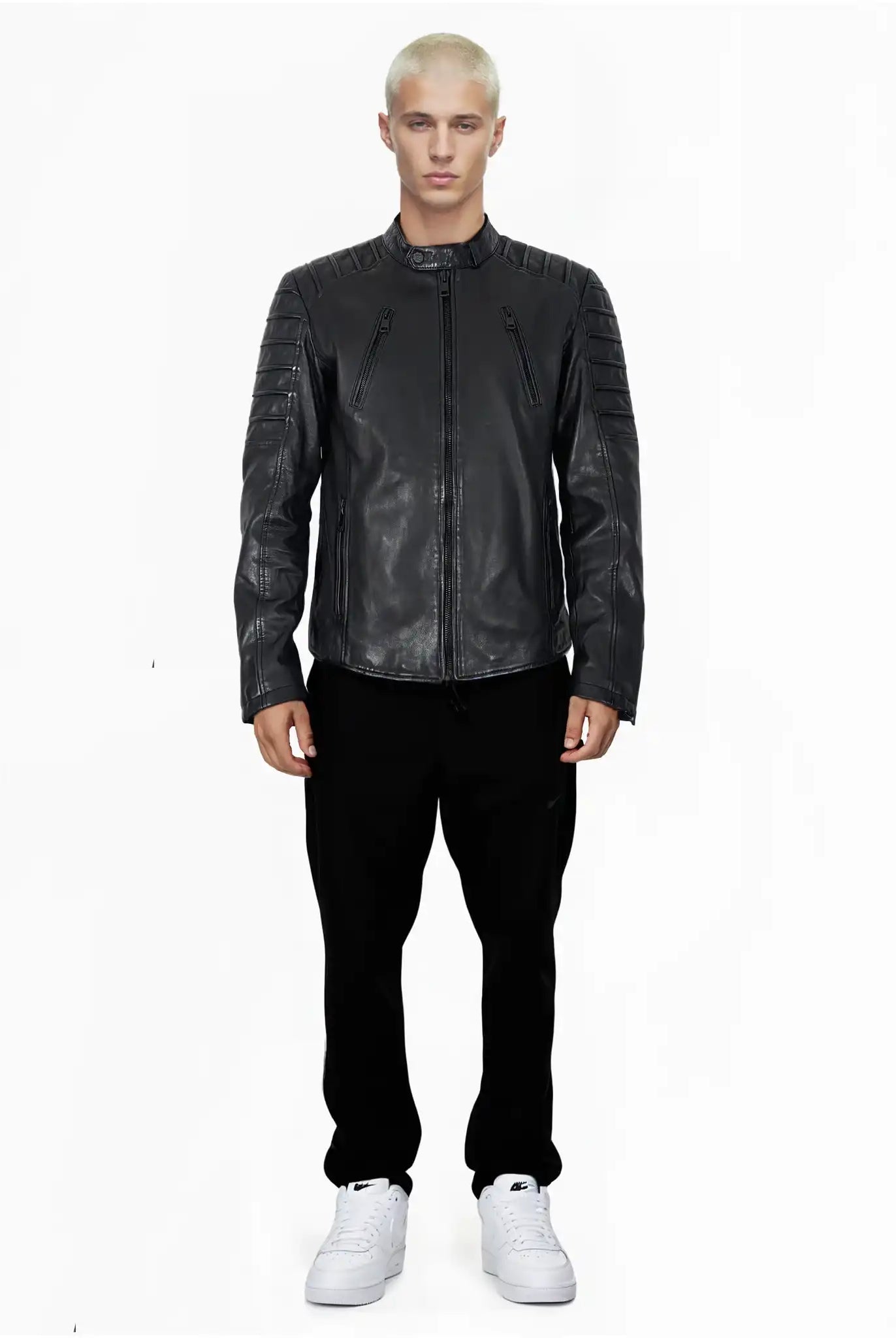 Eine Person mit kurzen, gebleichten blonden Haaren blickt nach vorne und trägt die trueprodigy Dash Slim-Fit-Lederjacke mit Stehkragen & Zipper-Details, eine schwarze Hose und weiße Turnschuhe vor einem schlichten weißen Hintergrund.