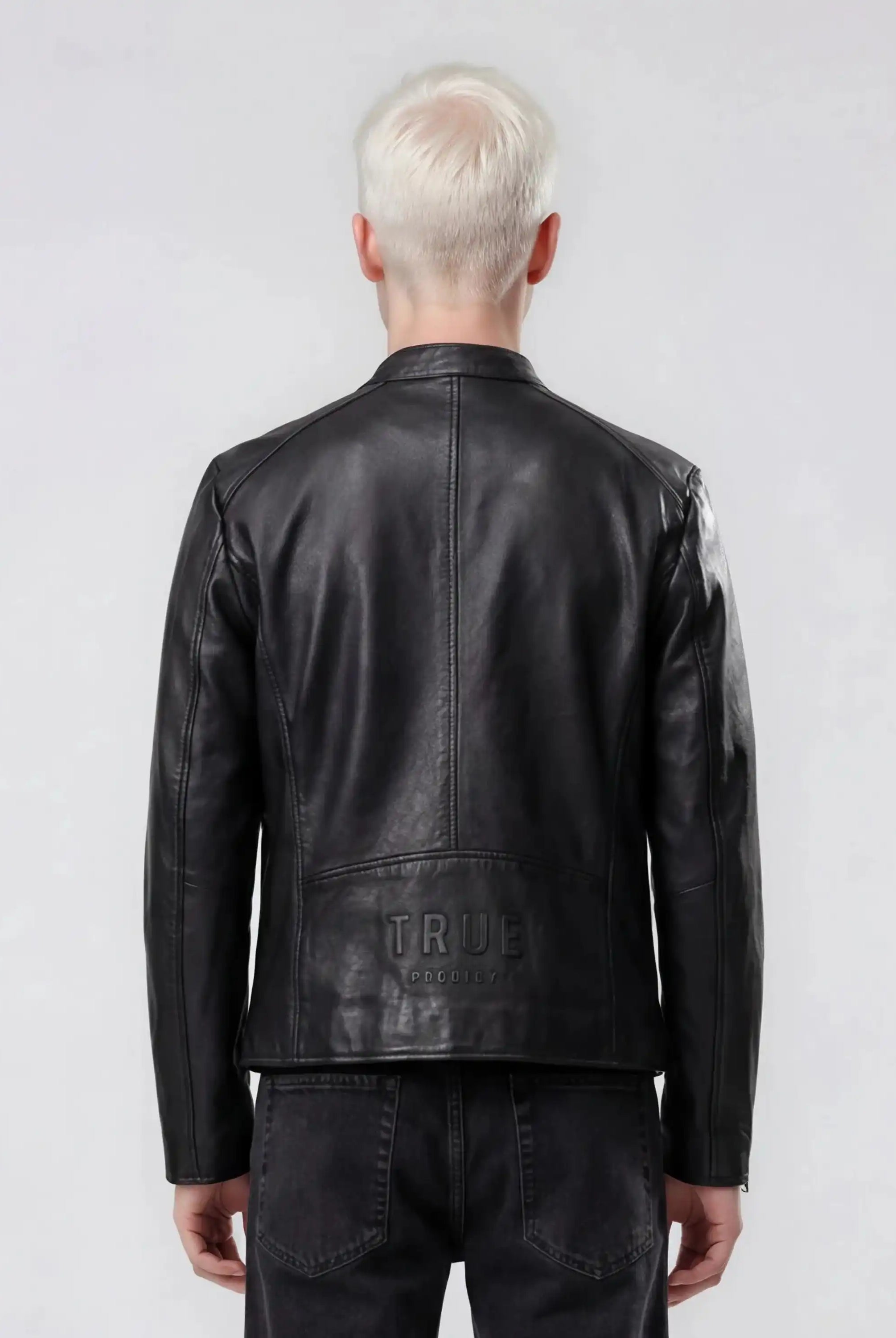 Eine Person mit kurzem platinblondem Haar, von hinten gesehen in schwarzer Jeans und einer trueprodigy Slim-Fit-Lederjacke mit sichtbarem 3D-Rückenlogo aus Schafsleder. Das Wort TRUE ist auf dem unteren Rücken der Jacke geprägt. Der Hintergrund ist hellgrau.