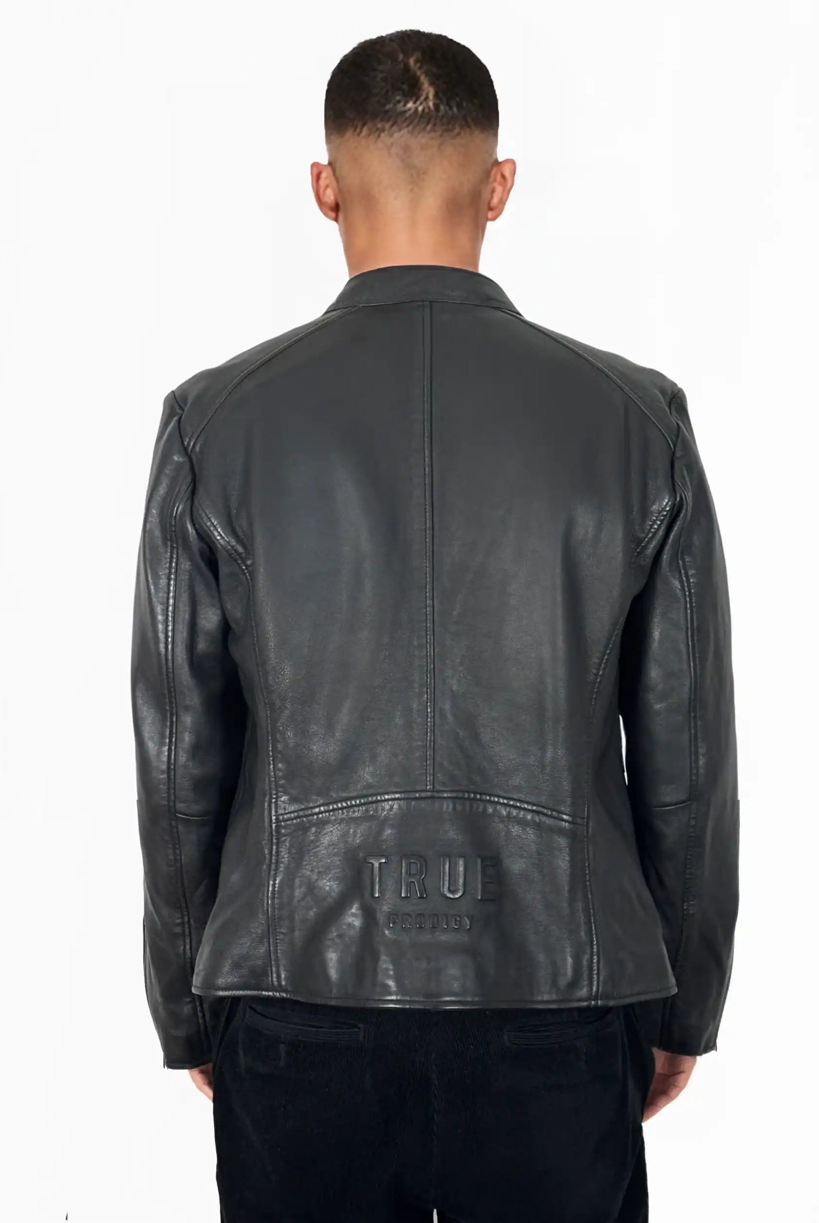 Eine Person schaut in einer Jonas Slim-Fit-Lederjacke mit sichtbarem 3D-Rückenlogo von trueprodigy, die über einer schwarzen Hose getragen wird, vor einem schlichten weißen Hintergrund von der Kamera weg.
