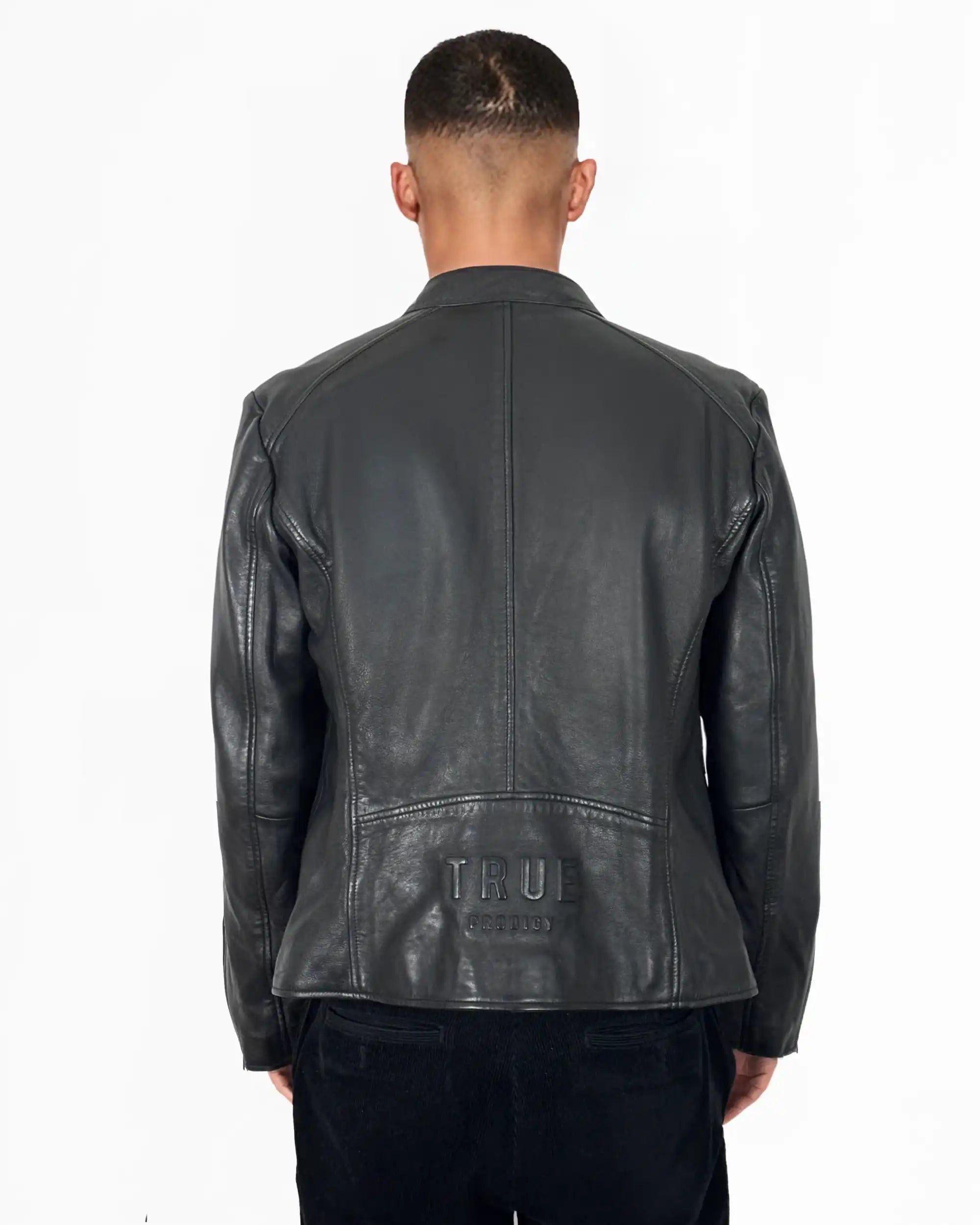 Eine Person schaut in einer Jonas Slim-Fit-Lederjacke mit sichtbarem 3D-Rückenlogo von trueprodigy, die über einer schwarzen Hose getragen wird, vor einem schlichten weißen Hintergrund von der Kamera weg.