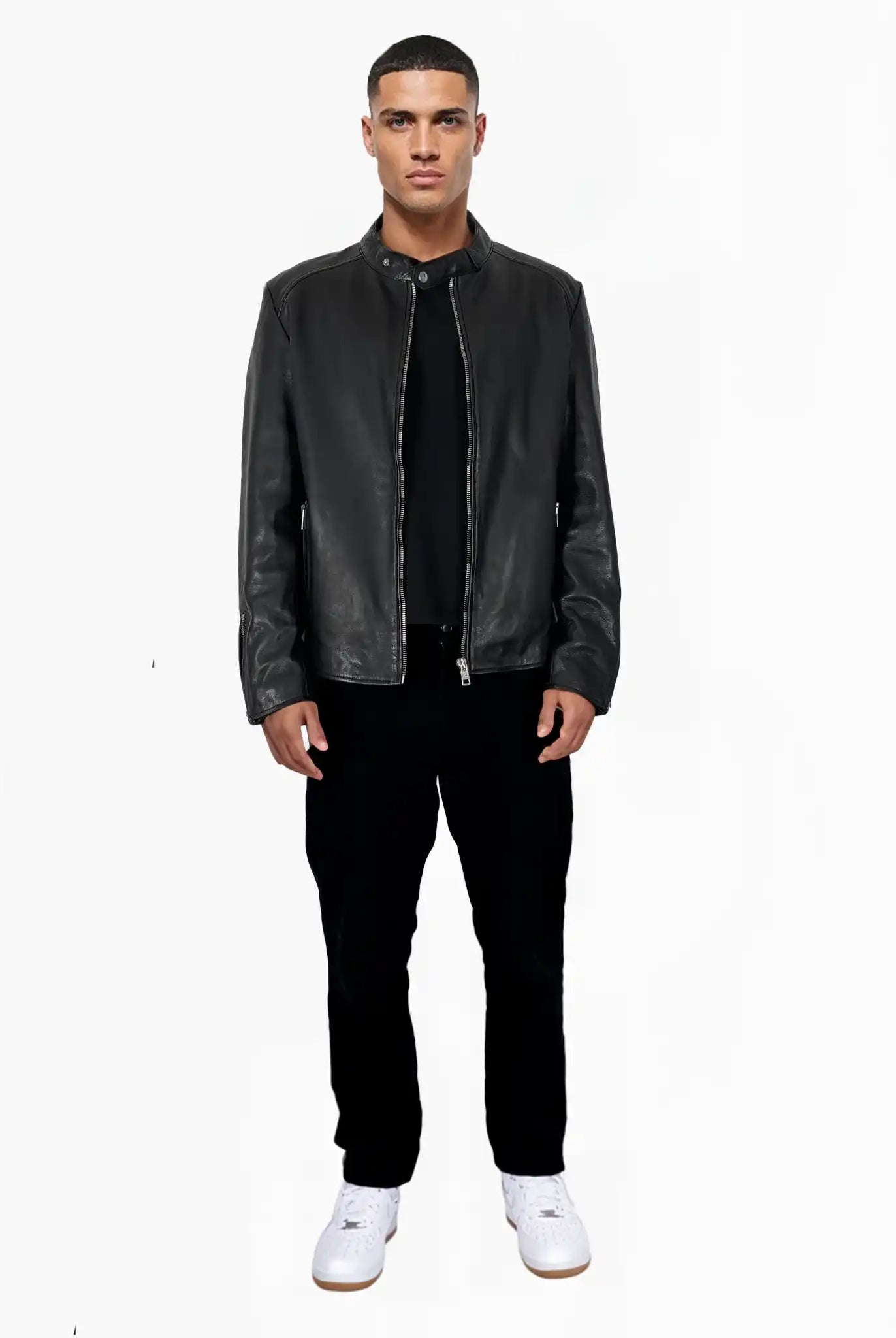 Ein Mann steht mit dem Gesicht nach vorne vor einem schlichten weißen Hintergrund und trägt die trueprodigy Jonas Slim-Fit-Lederjacke mit sichtbarem 3D-Rückenlogo, gepaart mit einem schwarzen Hemd, einer schwarzen Hose und weißen Sneakers.