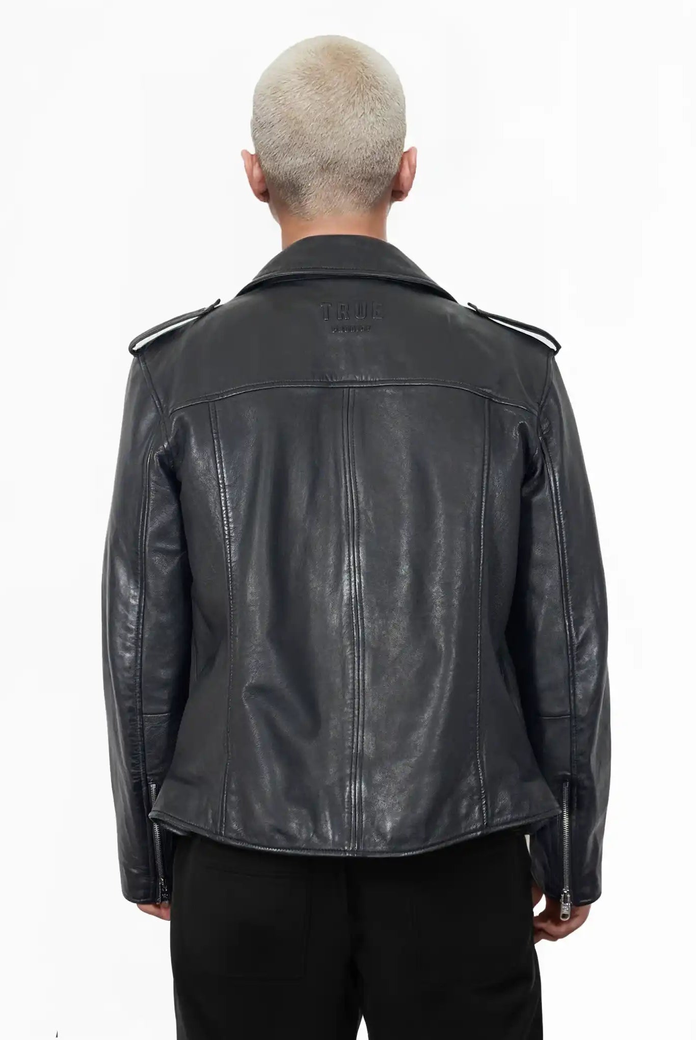 Eine Person mit kurzen, gebleichten Haaren ist von hinten zu sehen. Sie trägt die trueprodigy Easton Leder-Bikerjacke mit asymmetrischem Zipper & 3D-Logo unter dem Kragen und schwarzer Hose und setzt damit einen coolen Biker-Look vor einem schlichten weißen Hintergrund um.