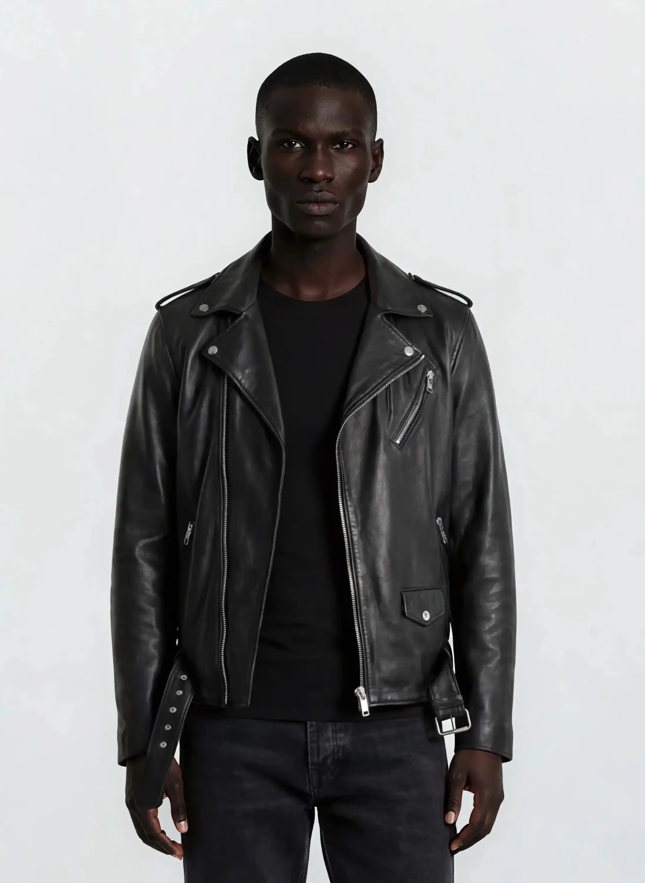 Eine Person blickt in die Kamera und trägt eine trueprodigy Leder-Bikerjacke mit asymmetrischem Zipper & 3D-Logo unter dem Kragen über einem schwarzen Hemd und einer dunklen Jeans vor einem schlichten hellgrauen Hintergrund.