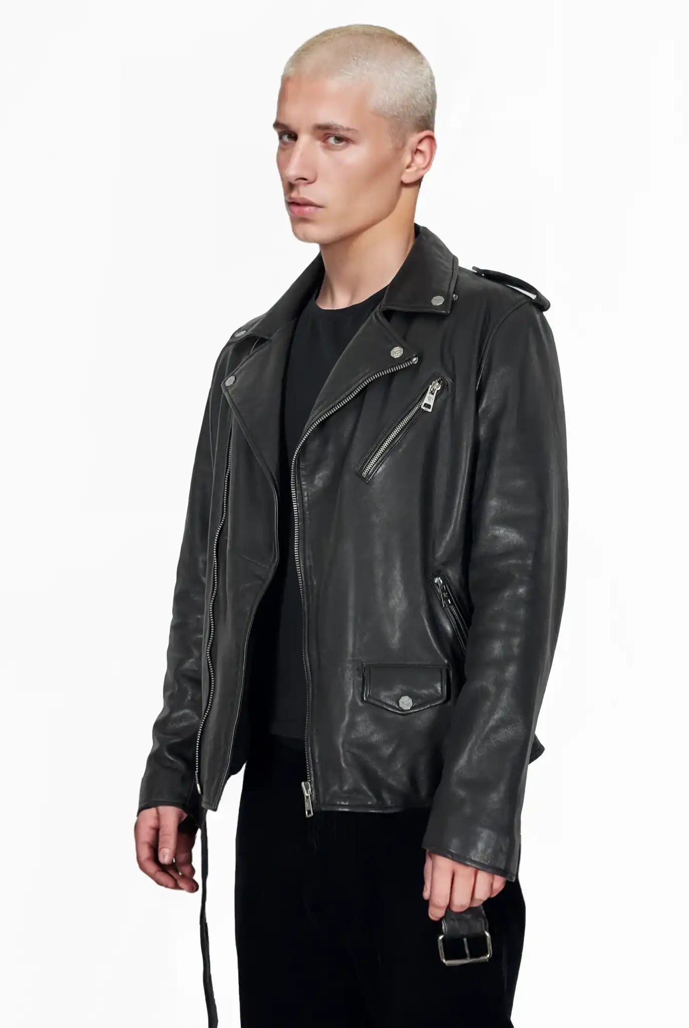 Eine Person mit kurzen, gebleichten blonden Haaren trägt eine trueprodigy Easton Leder-Bikerjacke mit asymmetrischem Zipper & 3D-Logo unter dem Kragen über einem schwarzen Hemd und einer Hose, stehend vor einem schlichten weißen Hintergrund.