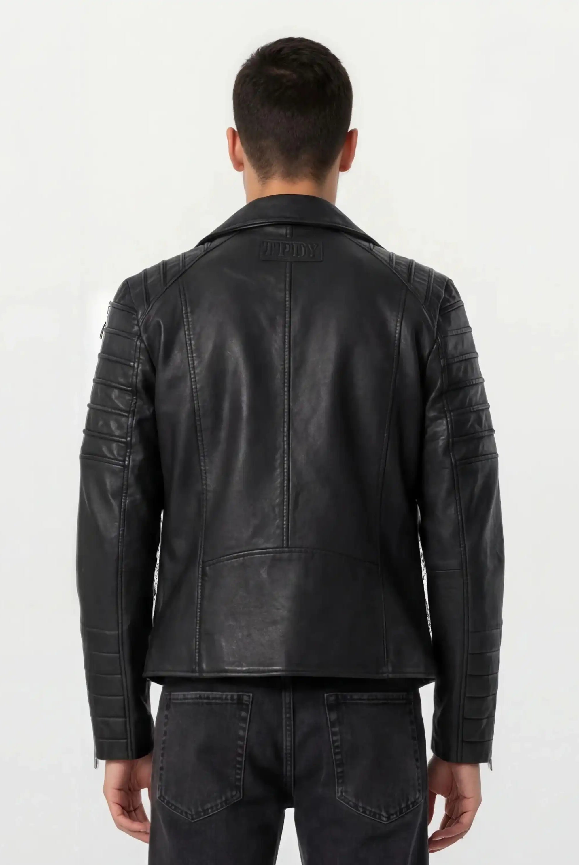 Ein Mann ist von hinten zu sehen, der eine Trueprodigy Lederjacke mit Paspelierung und diversen Reißverschlüssen trägt, gepaart mit einer dunklen Jeans vor einem schlichten hellen Hintergrund.