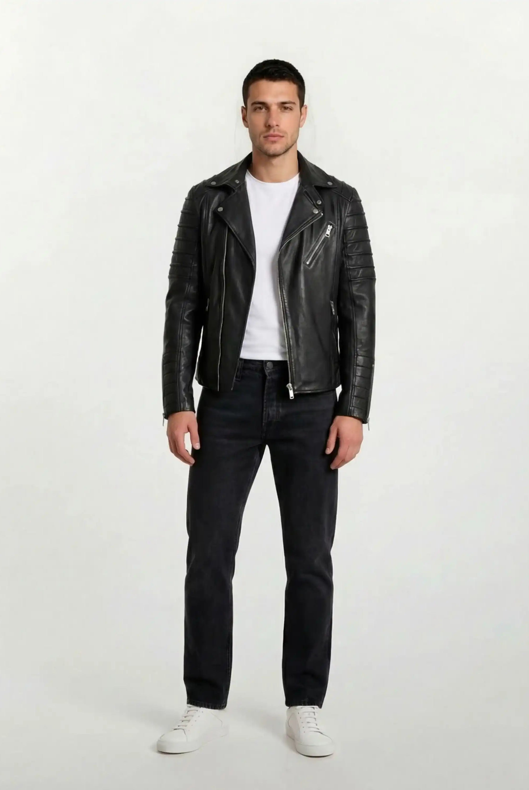 Ein Mann blickt nach vorne und trägt die trueprodigy Lederjacke mit Paspelierung und diversen Reißverschlüssen über einem weißen T-Shirt, dazu dunkle Jeans und weiße Turnschuhe. Der Hintergrund ist einfarbig und hell.