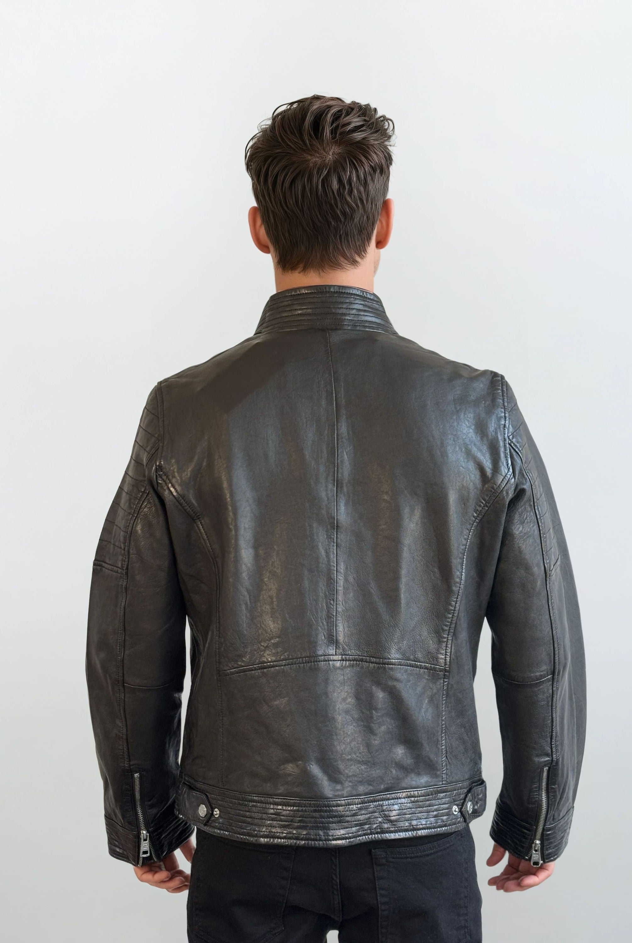 Eine Person mit kurzen braunen Haaren ist von hinten zu sehen, die eine trueprodigy Herren Lederjacke aus Schafsleder in Schwarz mit klassischem Blouson-Schnitt und eine schwarze Hose vor einem schlichten hellen Hintergrund trägt.