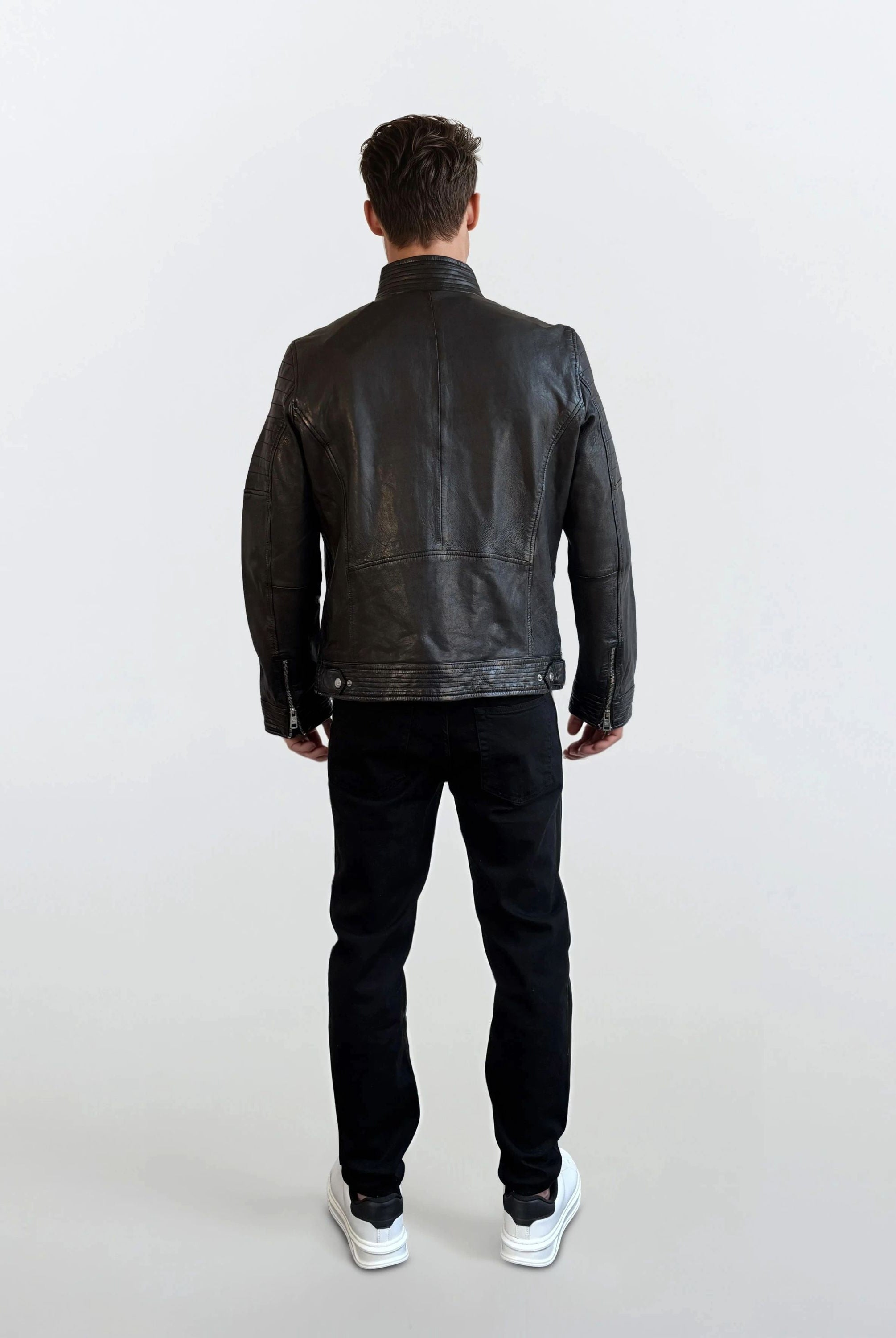 Eine Person steht mit dem Rücken zur Kamera vor hellem Hintergrund und trägt eine schwarze trueprodigy Herren Lederjacke aus Schafsleder im klassischen Blouson-Schnitt, dazu schwarze Hose und weiße Sneaker.