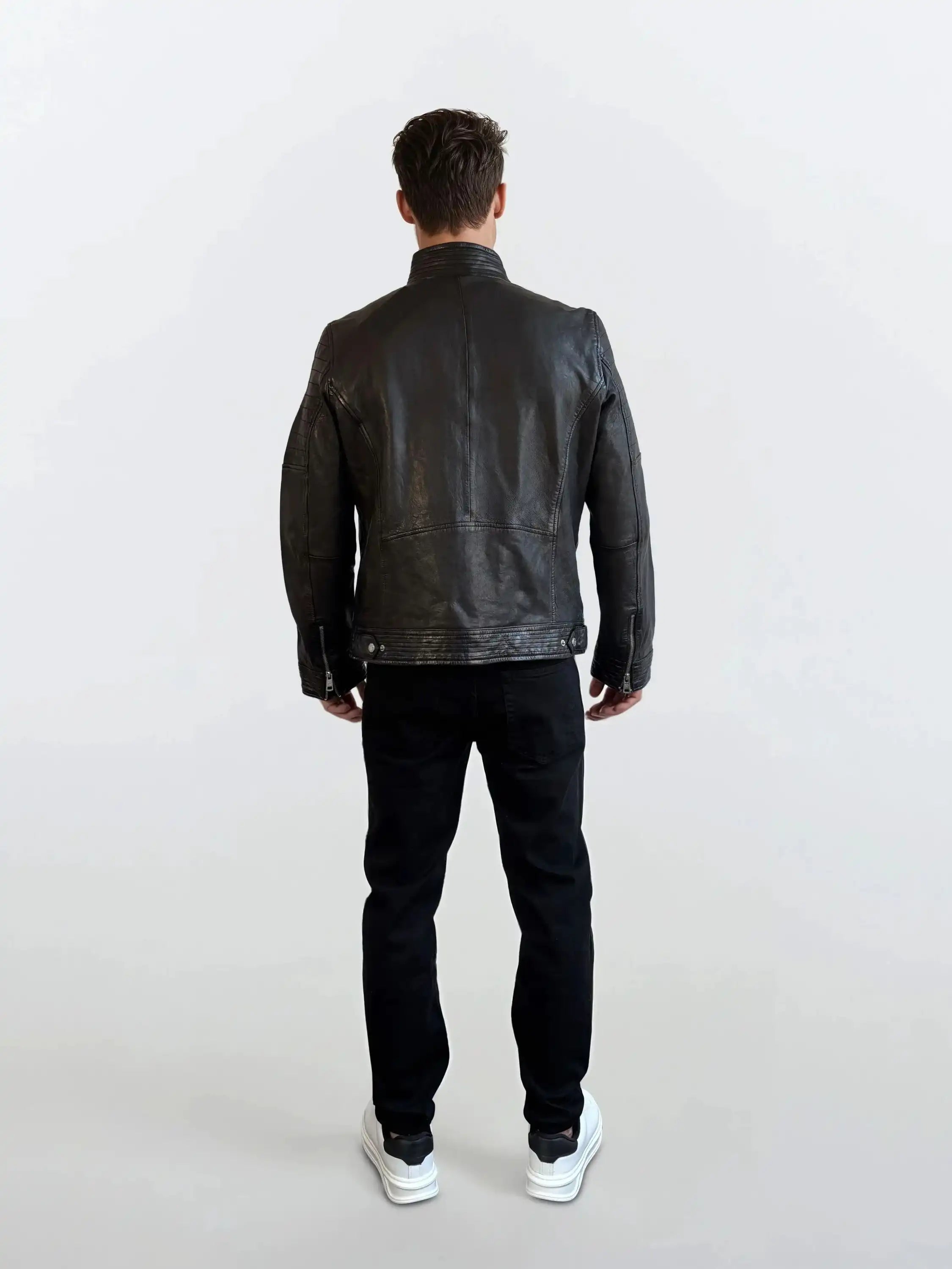 Eine Person steht mit dem Rücken zur Kamera vor hellem Hintergrund und trägt eine schwarze trueprodigy Herren Lederjacke aus Schafsleder im klassischen Blouson-Schnitt, dazu schwarze Hose und weiße Sneaker.