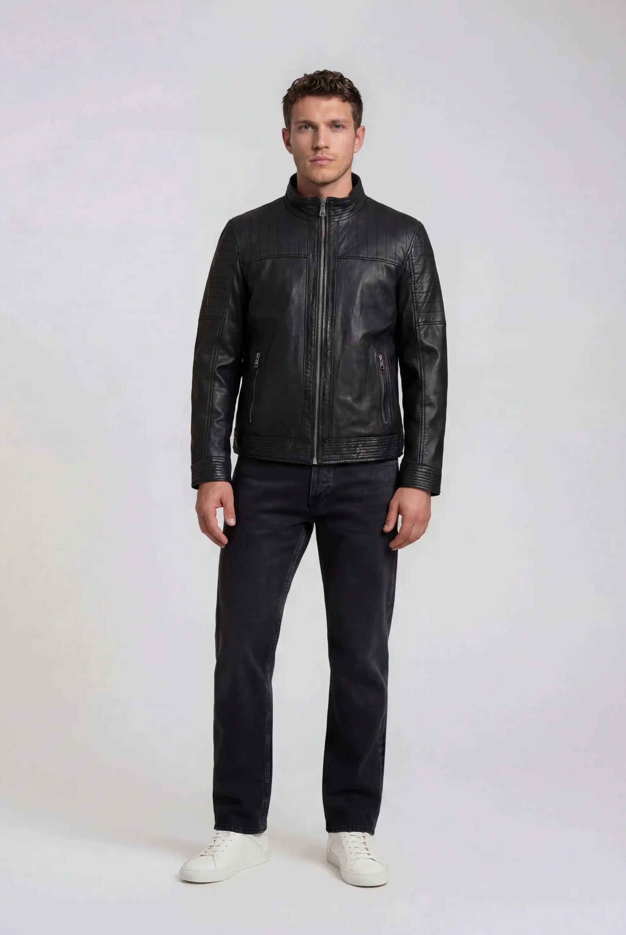 Ein Mann trägt die schwarze trueprodigy Herren Lederjacke aus Schafsleder im klassischen Blouson-Schnitt, kombiniert mit dunkler Jeans und weißen Sneakers. Seine Arme hängen locker an den Seiten, während er vor einem hellen Hintergrund steht.