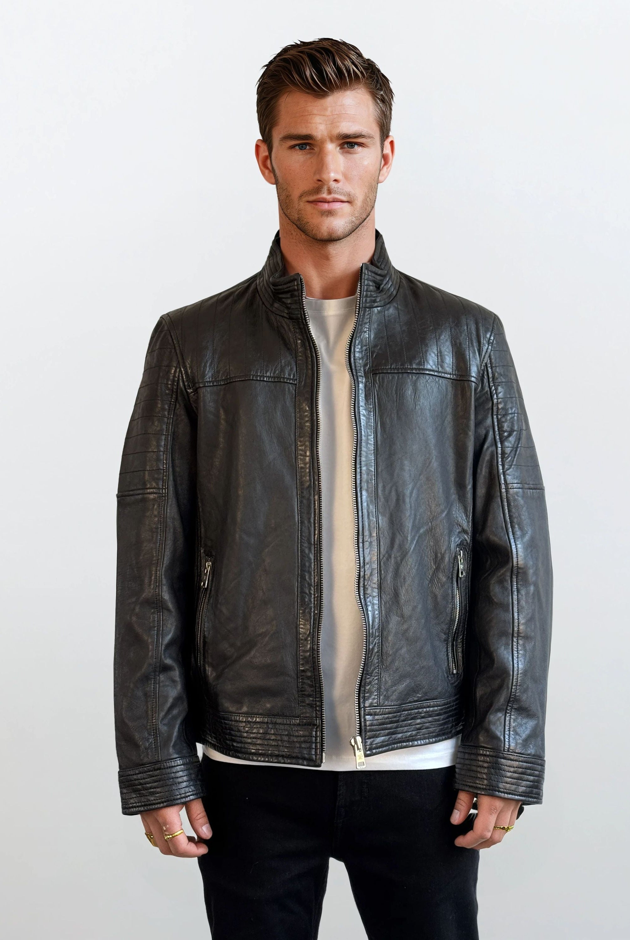 Ein Mann mit kurzen braunen Haaren steht mit dem Gesicht nach vorne vor einem schlichten hellen Hintergrund und trägt eine schwarze Herren Lederjacke aus Schafsleder im klassischen Blouson-Schnitt von trueprodigy über einem weißen Shirt und schwarzen Hosen. Seine Hände liegen entspannt an den Seiten.