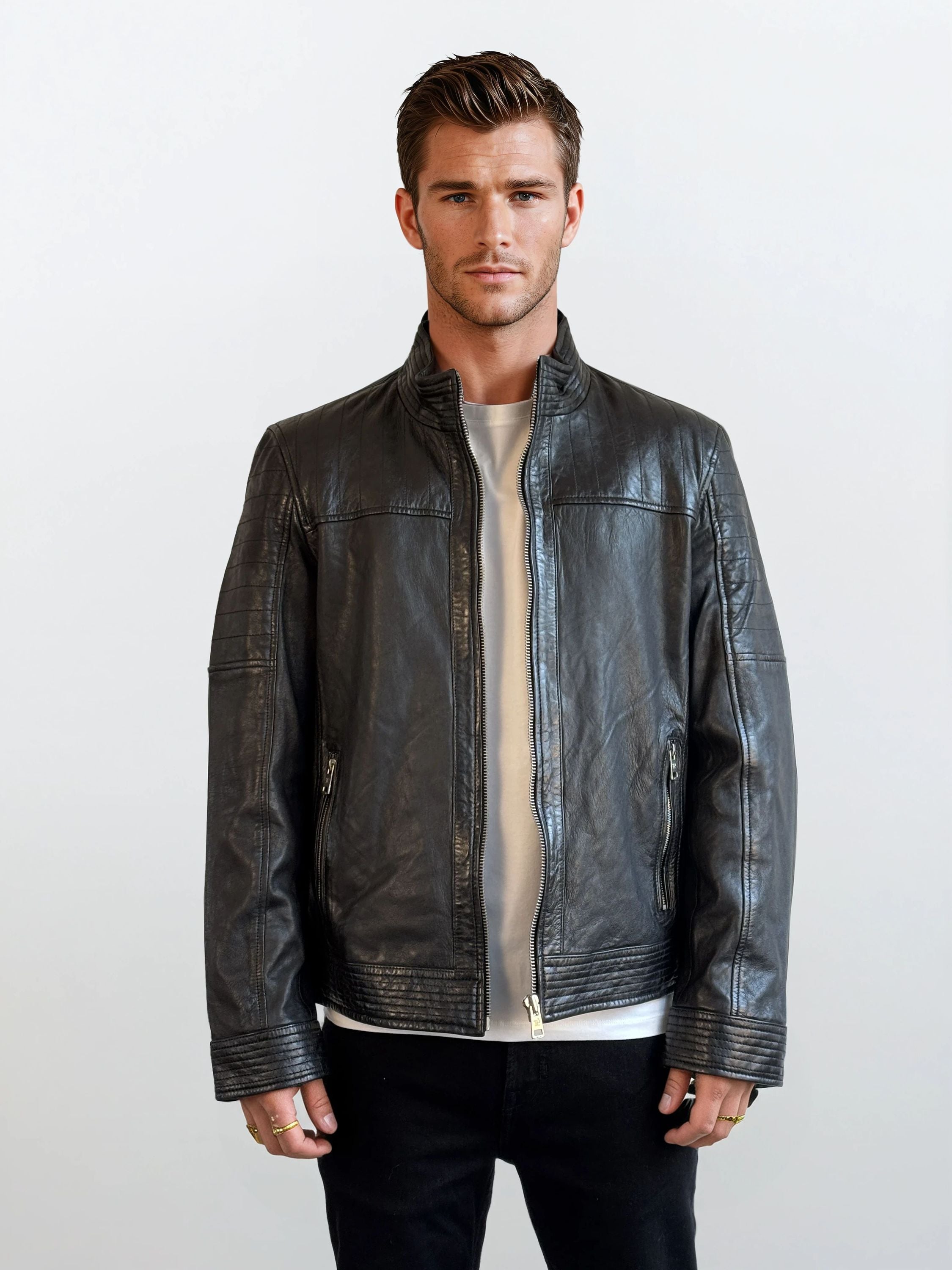 Ein Mann mit kurzen braunen Haaren steht mit dem Gesicht nach vorne vor einem schlichten hellen Hintergrund und trägt eine schwarze Herren Lederjacke aus Schafsleder im klassischen Blouson-Schnitt von trueprodigy über einem weißen Shirt und schwarzen Hosen. Seine Hände liegen entspannt an den Seiten.