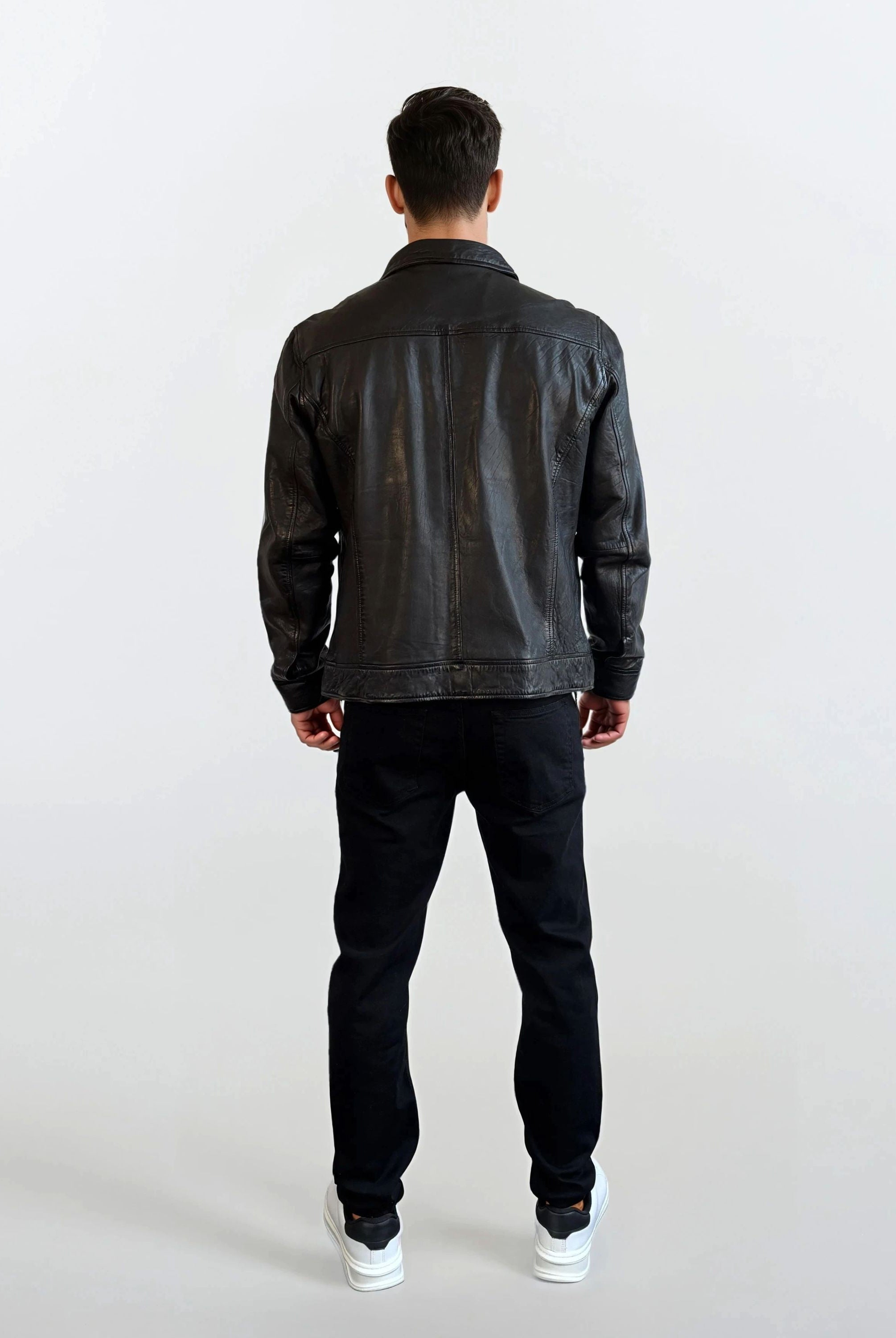 Eine Person trägt die Herren Lederjacke aus Schafsleder in Schwarz mit Hemd-Design von trueprodigy, dazu schwarze Hosen und weiße Sneakers, und steht vor einem schlichten hellen Hintergrund mit dem Rücken zur Kamera.