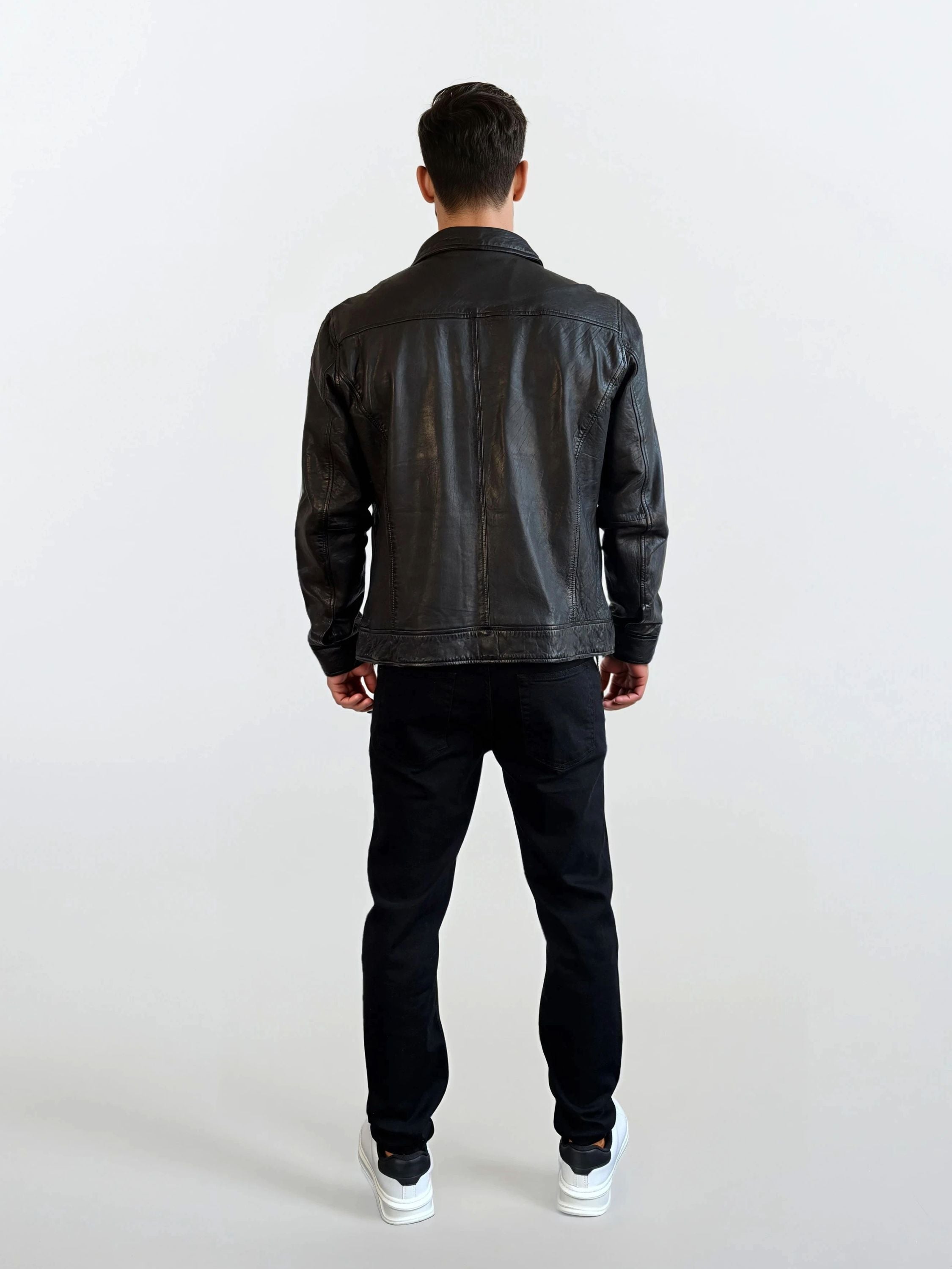 Eine Person trägt die Herren Lederjacke aus Schafsleder in Schwarz mit Hemd-Design von trueprodigy, dazu schwarze Hosen und weiße Sneakers, und steht vor einem schlichten hellen Hintergrund mit dem Rücken zur Kamera.