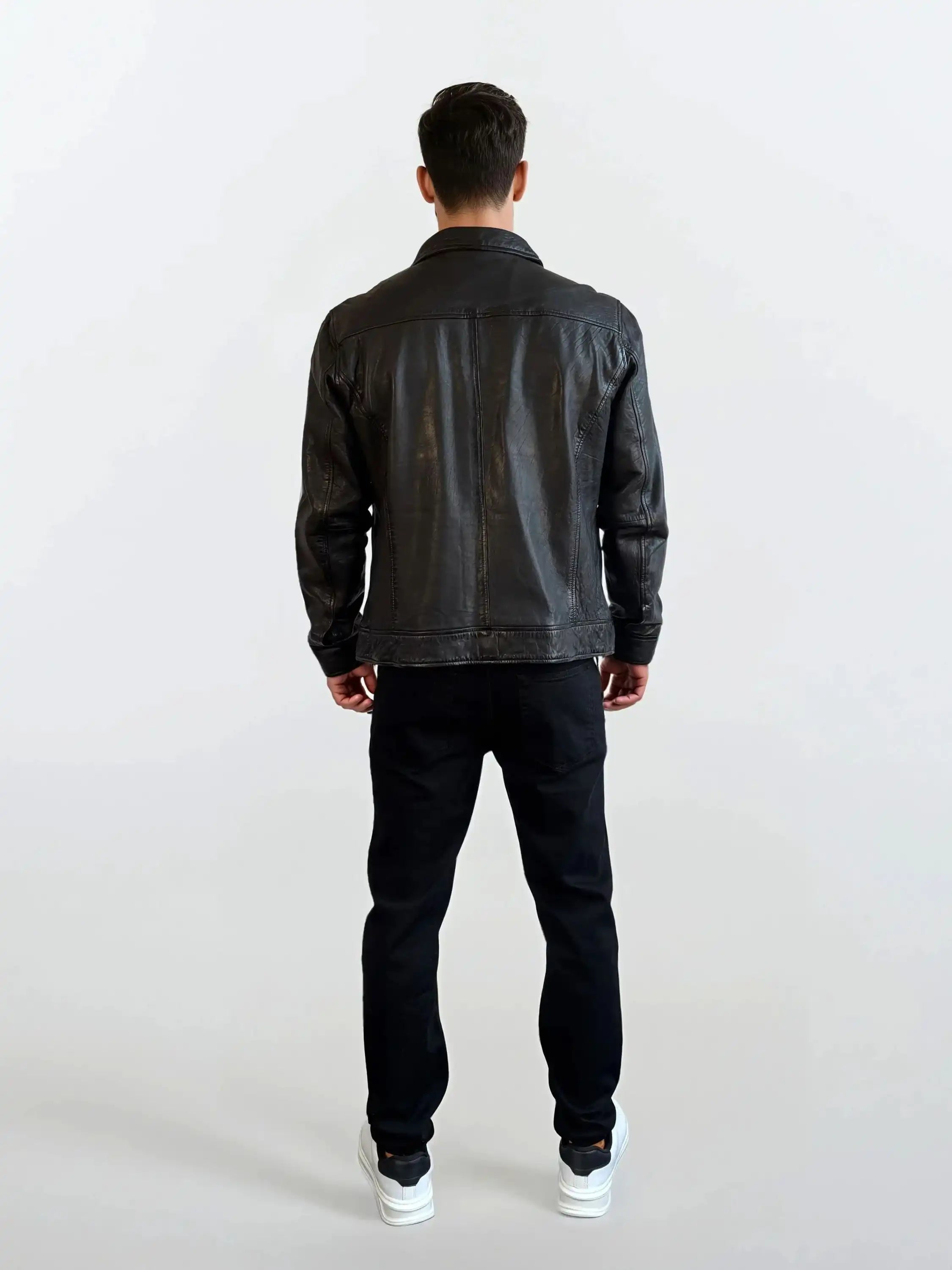 Eine Person trägt die Herren Lederjacke aus Schafsleder in Schwarz mit Hemd-Design von trueprodigy, dazu schwarze Hosen und weiße Sneakers, und steht vor einem schlichten hellen Hintergrund mit dem Rücken zur Kamera.