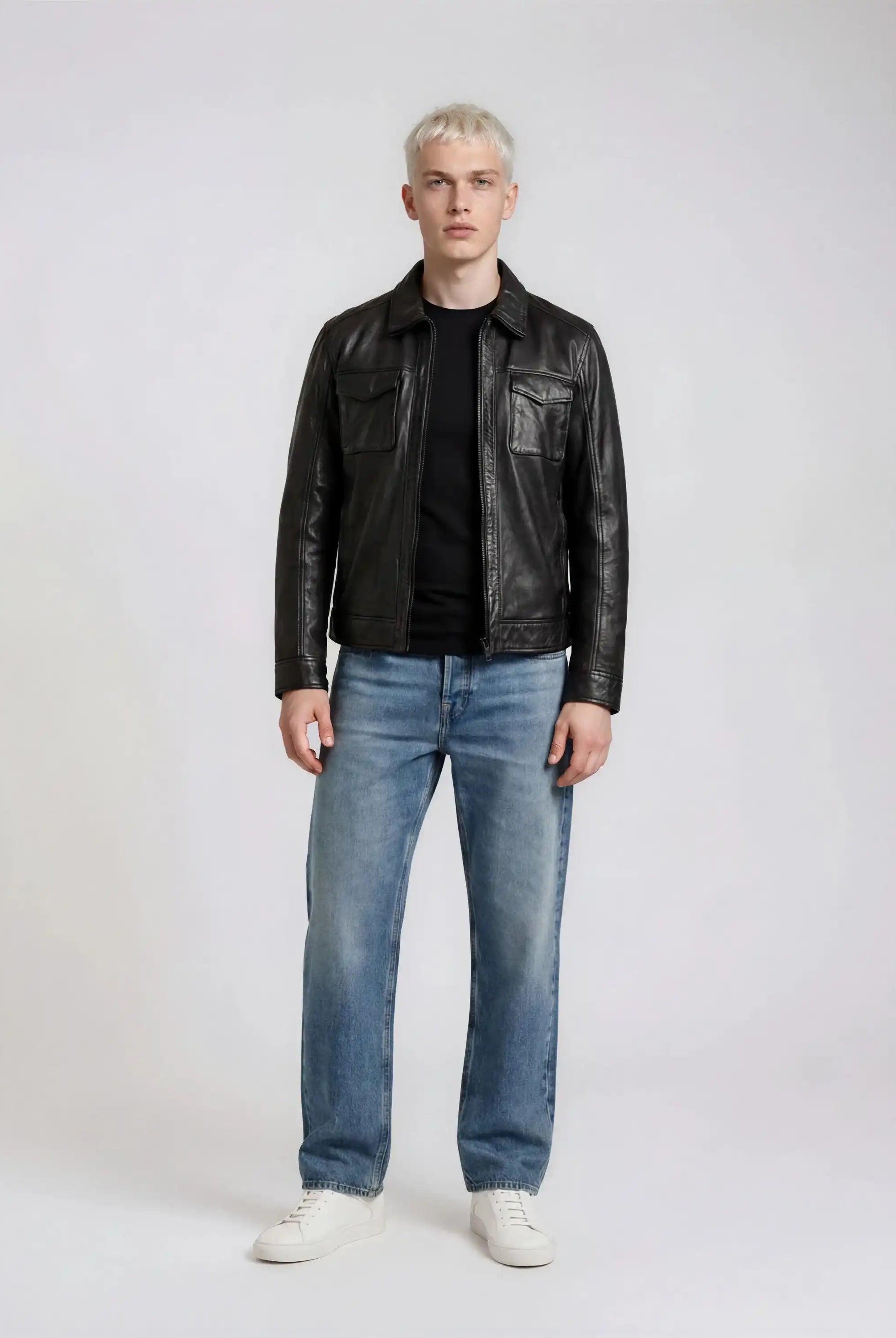 Eine Person mit kurzen blonden Haaren steht mit dem Gesicht nach vorne und trägt eine trueprodigy Herren Lederjacke aus Schafsleder in Schwarz mit Hemd-Design, gepaart mit einem schwarzen Hemd, blauen Jeans und weißen Turnschuhen vor einem schlichten hellen Hintergrund.