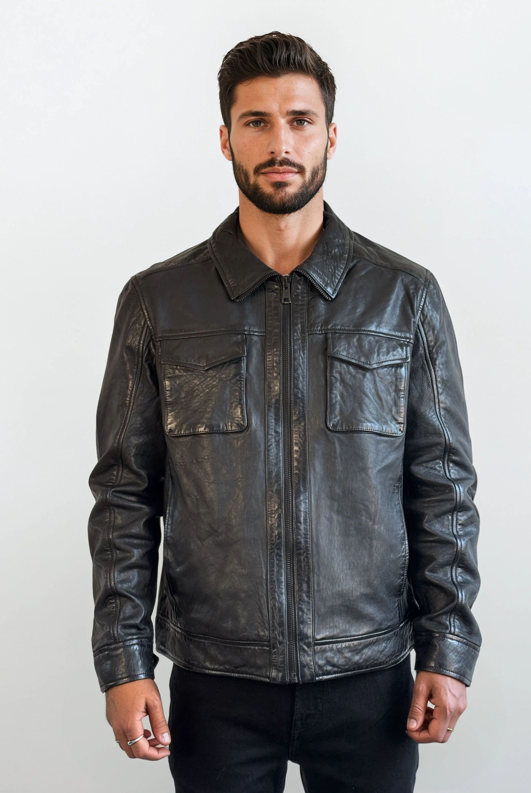 Ein Mann mit kurzen dunklen Haaren und Bart steht vor einem schlichten weißen Hintergrund, trägt die trueprodigy Herren Lederjacke aus Schafsleder in Schwarz mit Hemd-Design und eine schwarze Hose und blickt mit neutralem Blick in die Kamera.
