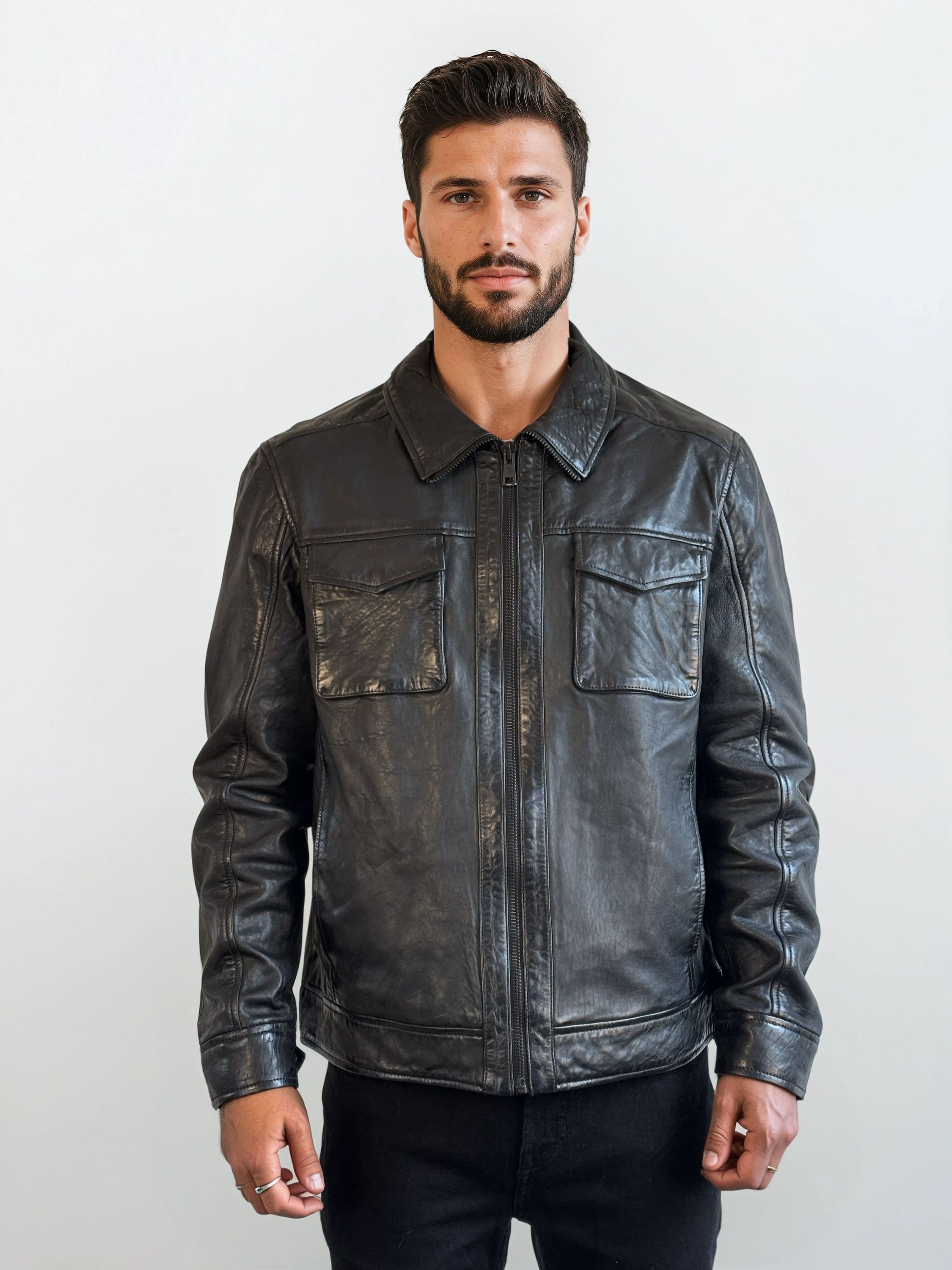 Ein Mann mit kurzen dunklen Haaren und Bart steht vor einem schlichten weißen Hintergrund, trägt die trueprodigy Herren Lederjacke aus Schafsleder in Schwarz mit Hemd-Design und eine schwarze Hose und blickt mit neutralem Blick in die Kamera.