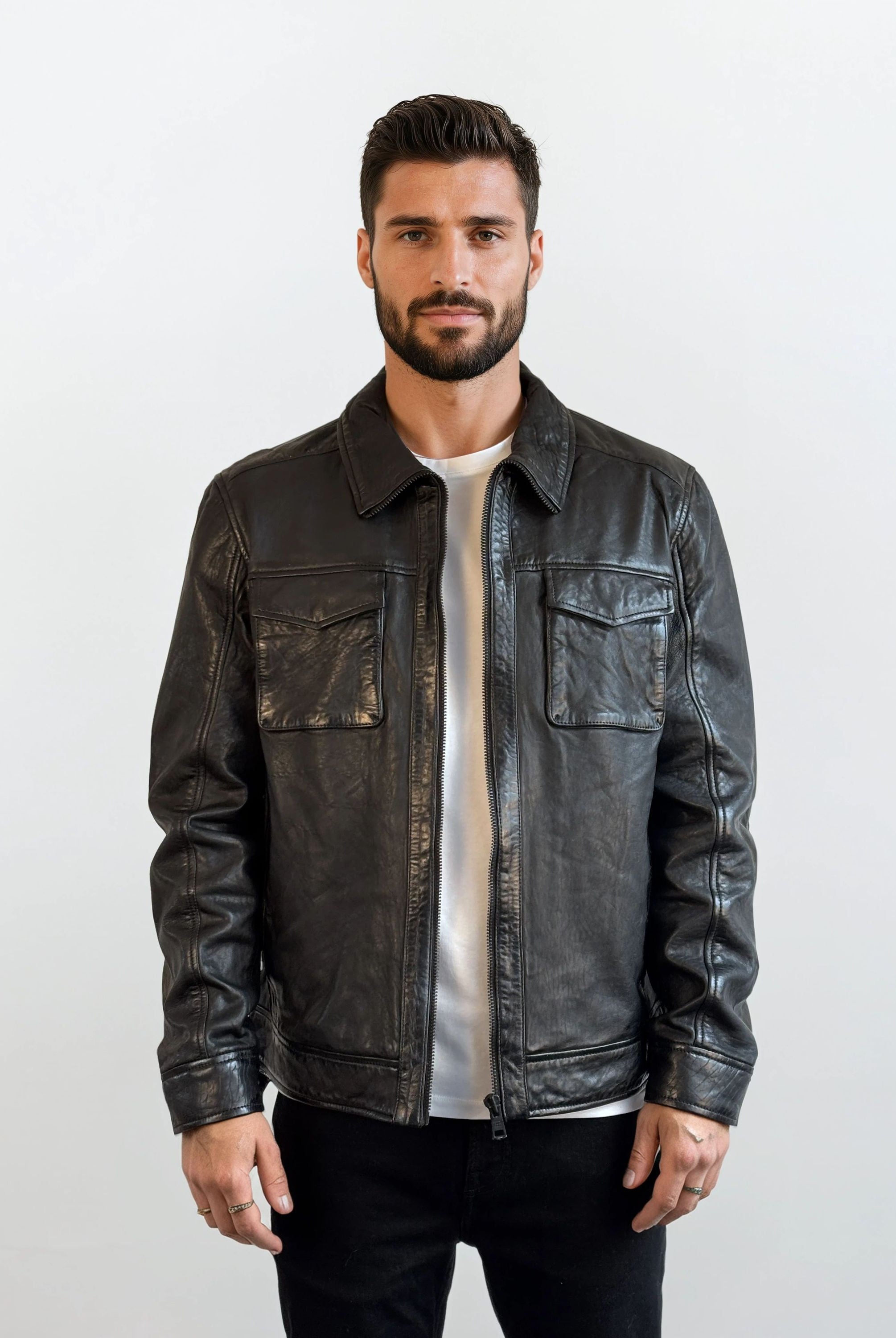 Ein Mann mit kurzen dunklen Haaren und Bart steht vor einem hellen Hintergrund, trägt die trueprodigy Herren Lederjacke aus Schafsleder in Schwarz mit Hemd-Design über einem weißen Hemd und einer schwarzen Hose und schaut neutral in die Kamera.