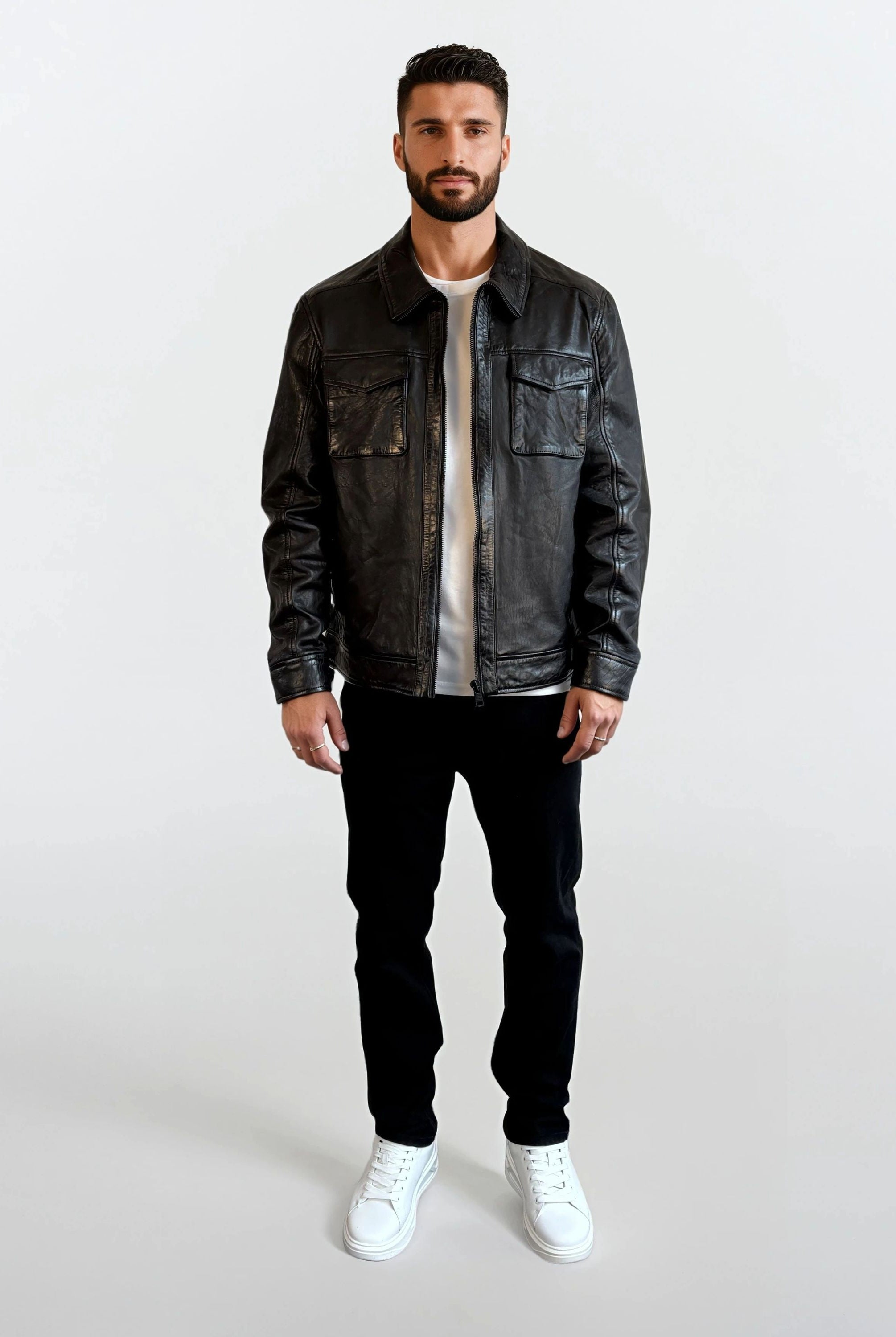 Ein Mann mit kurzem dunklem Haar und Bart trägt die Herren Lederjacke aus Schafsleder in Schwarz mit Hemd-Design von trueprodigy, dazu weißes Shirt, schwarze Hose und weiße Sneakers vor schlichtem weißem Hintergrund.