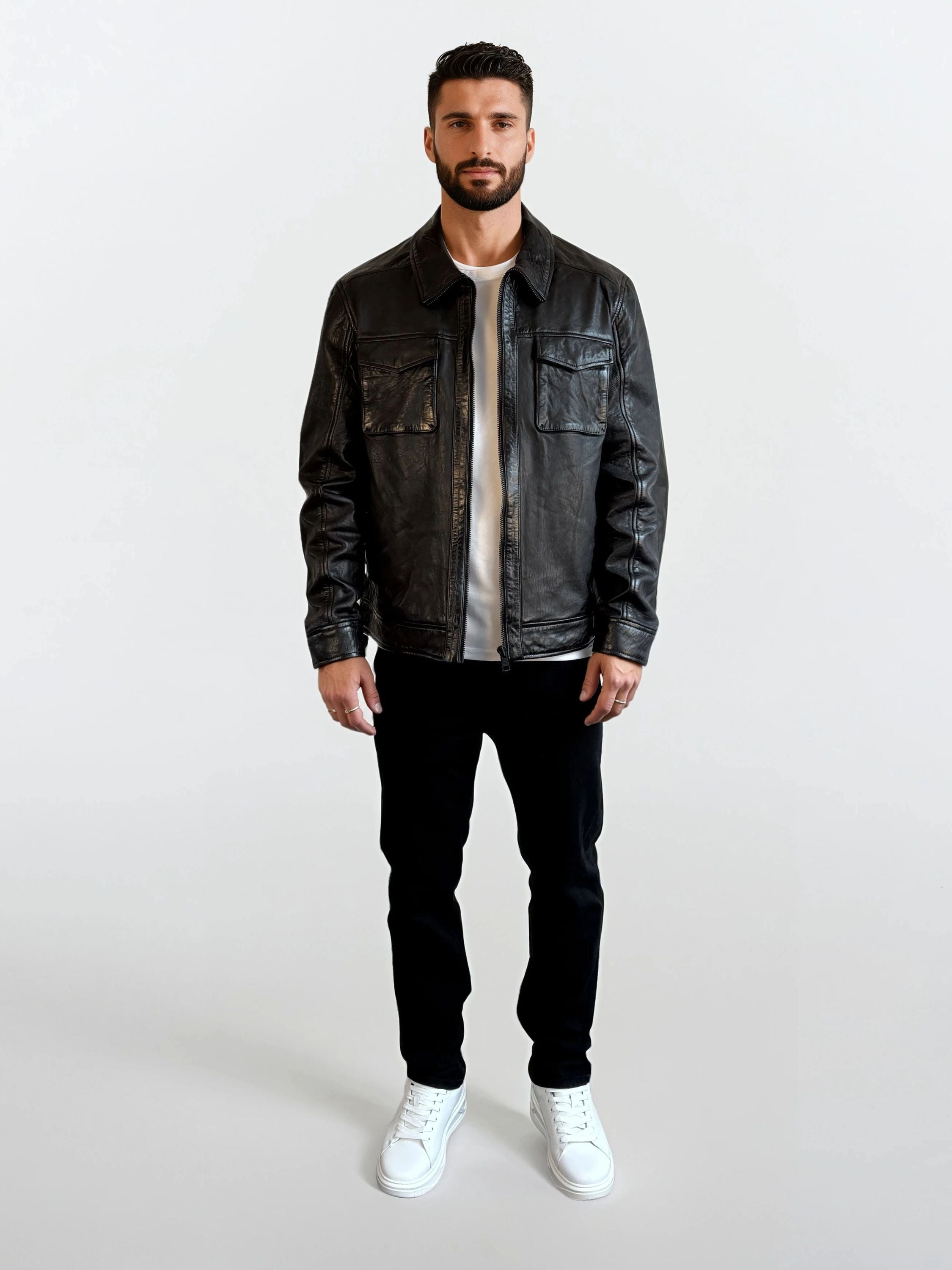 Ein Mann mit kurzem dunklem Haar und Bart trägt die Herren Lederjacke aus Schafsleder in Schwarz mit Hemd-Design von trueprodigy, dazu weißes Shirt, schwarze Hose und weiße Sneakers vor schlichtem weißem Hintergrund.