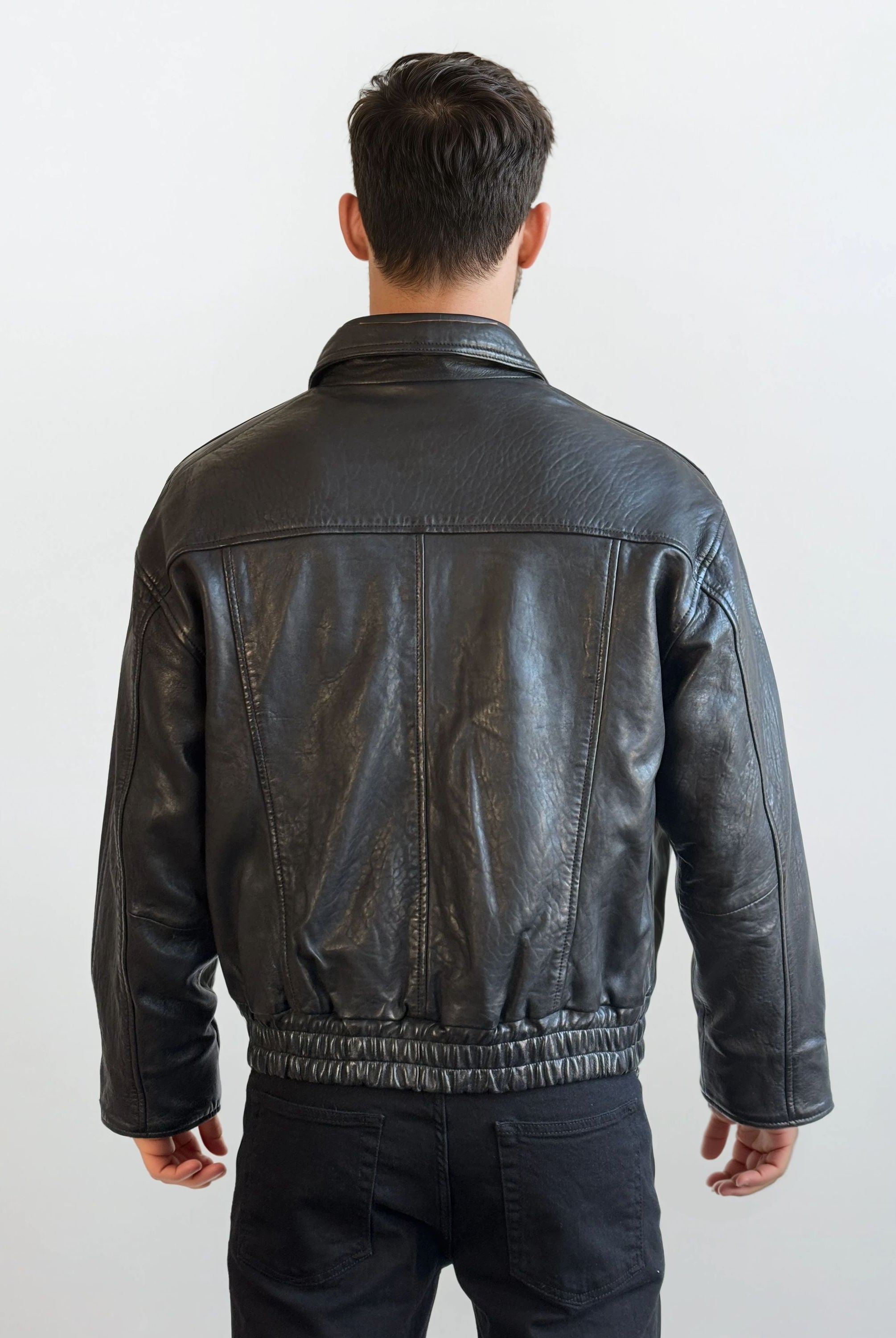 Eine Person mit kurzen braunen Haaren steht mit dem Rücken zur Kamera und trägt eine trueprodigy Herren Lederjacke aus Schafsleder in Schwarz mit klassischem Blouson-Schnitt und schwarzer Hose vor einem schlichten hellen Hintergrund.