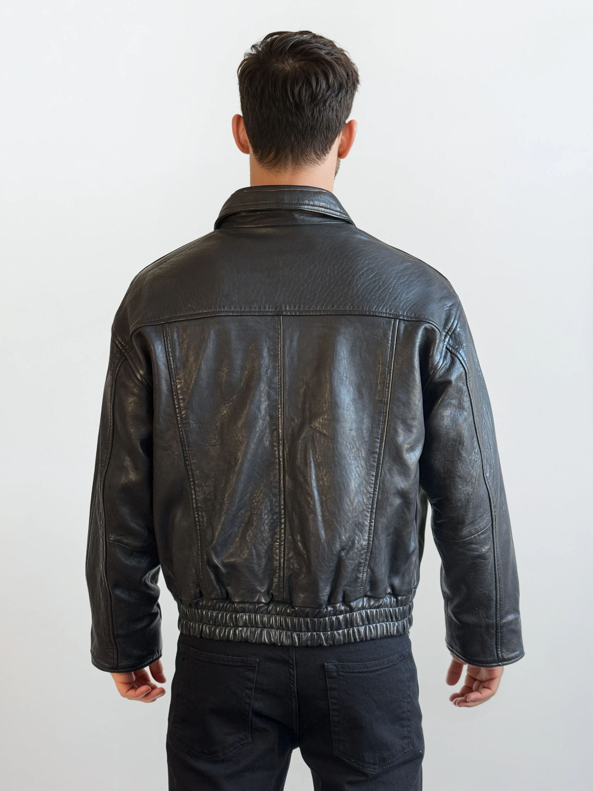 Eine Person mit kurzen braunen Haaren steht mit dem Rücken zur Kamera und trägt eine trueprodigy Herren Lederjacke aus Schafsleder in Schwarz mit klassischem Blouson-Schnitt und schwarzer Hose vor einem schlichten hellen Hintergrund.