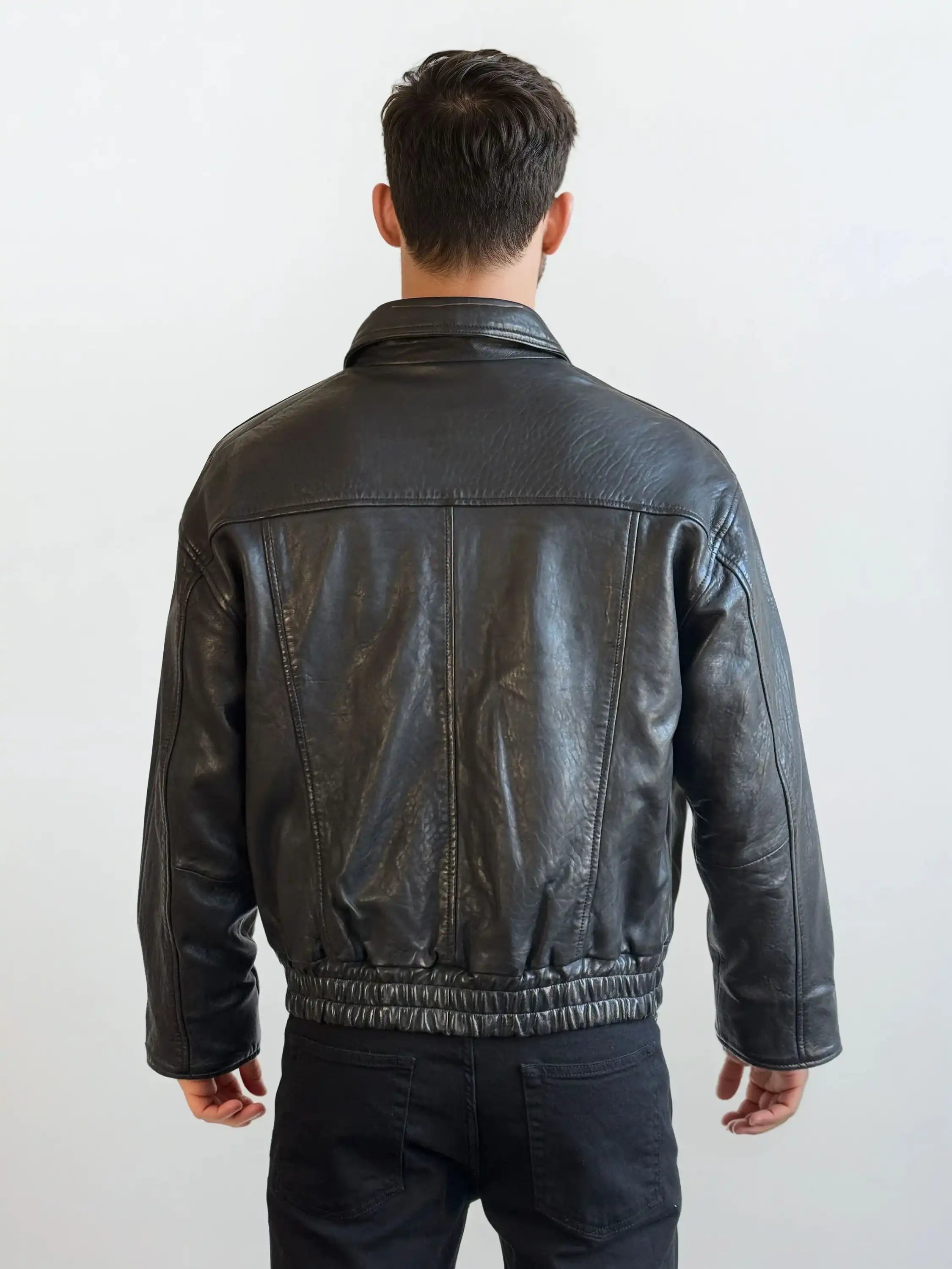 Eine Person mit kurzen braunen Haaren steht mit dem Rücken zur Kamera und trägt eine trueprodigy Herren Lederjacke aus Schafsleder in Schwarz mit klassischem Blouson-Schnitt und schwarzer Hose vor einem schlichten hellen Hintergrund.