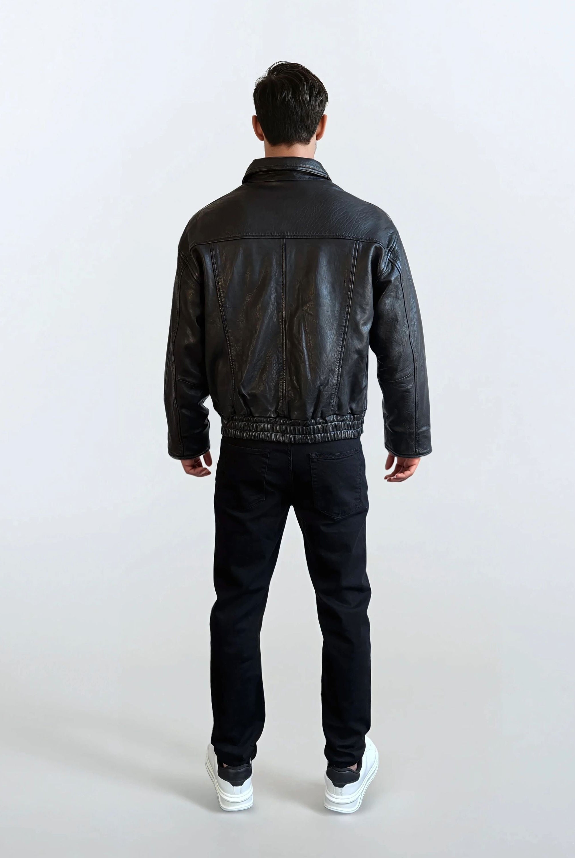 Eine Person blickt von der Kamera weg und trägt die trueprodigy Herren Lederjacke aus Schafsleder in Schwarz mit klassischem Blouson-Schnitt, eine schwarze Hose und weiße Turnschuhe vor einem schlichten hellen Hintergrund.