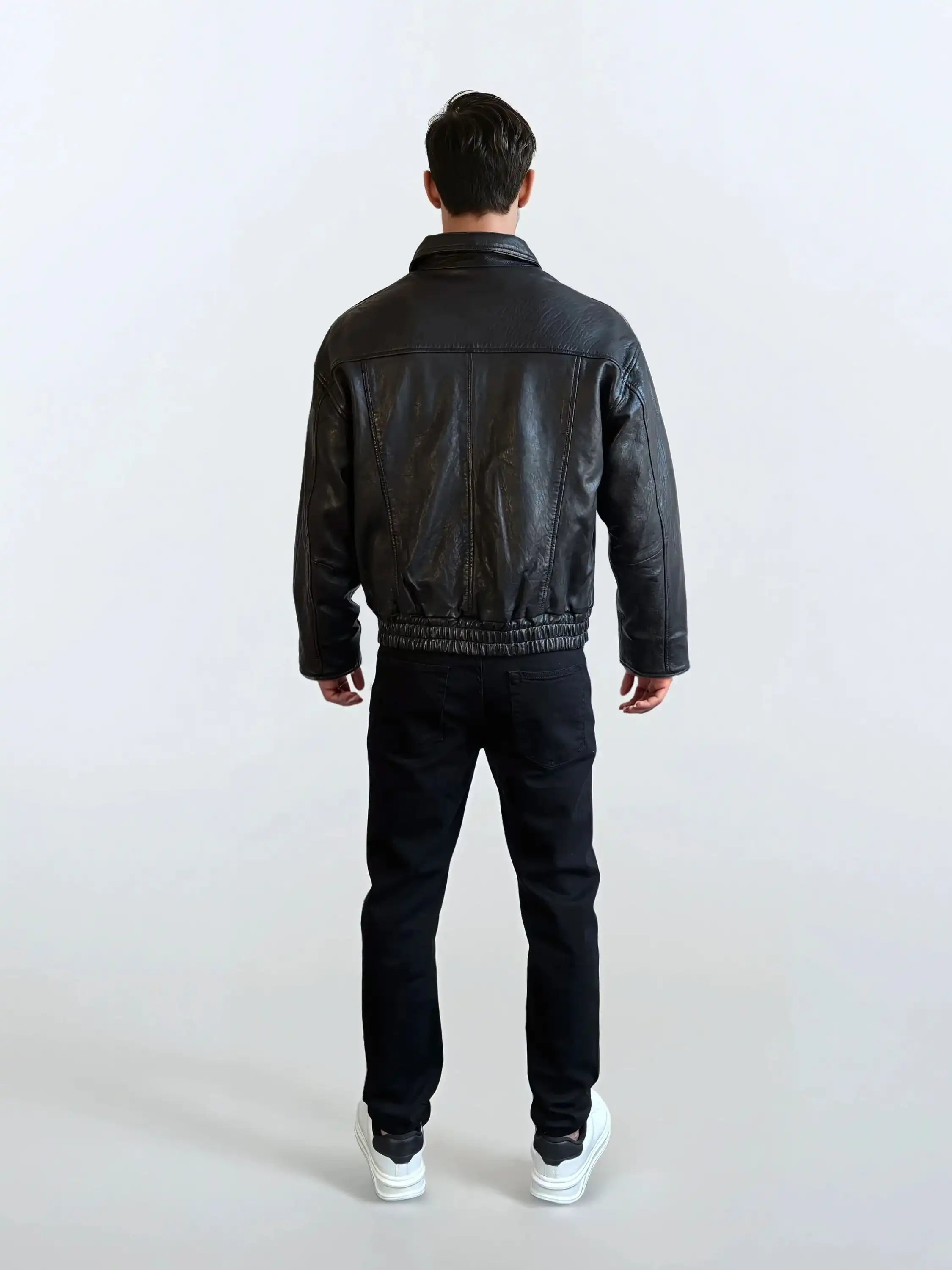 Eine Person blickt von der Kamera weg und trägt die trueprodigy Herren Lederjacke aus Schafsleder in Schwarz mit klassischem Blouson-Schnitt, eine schwarze Hose und weiße Turnschuhe vor einem schlichten hellen Hintergrund.