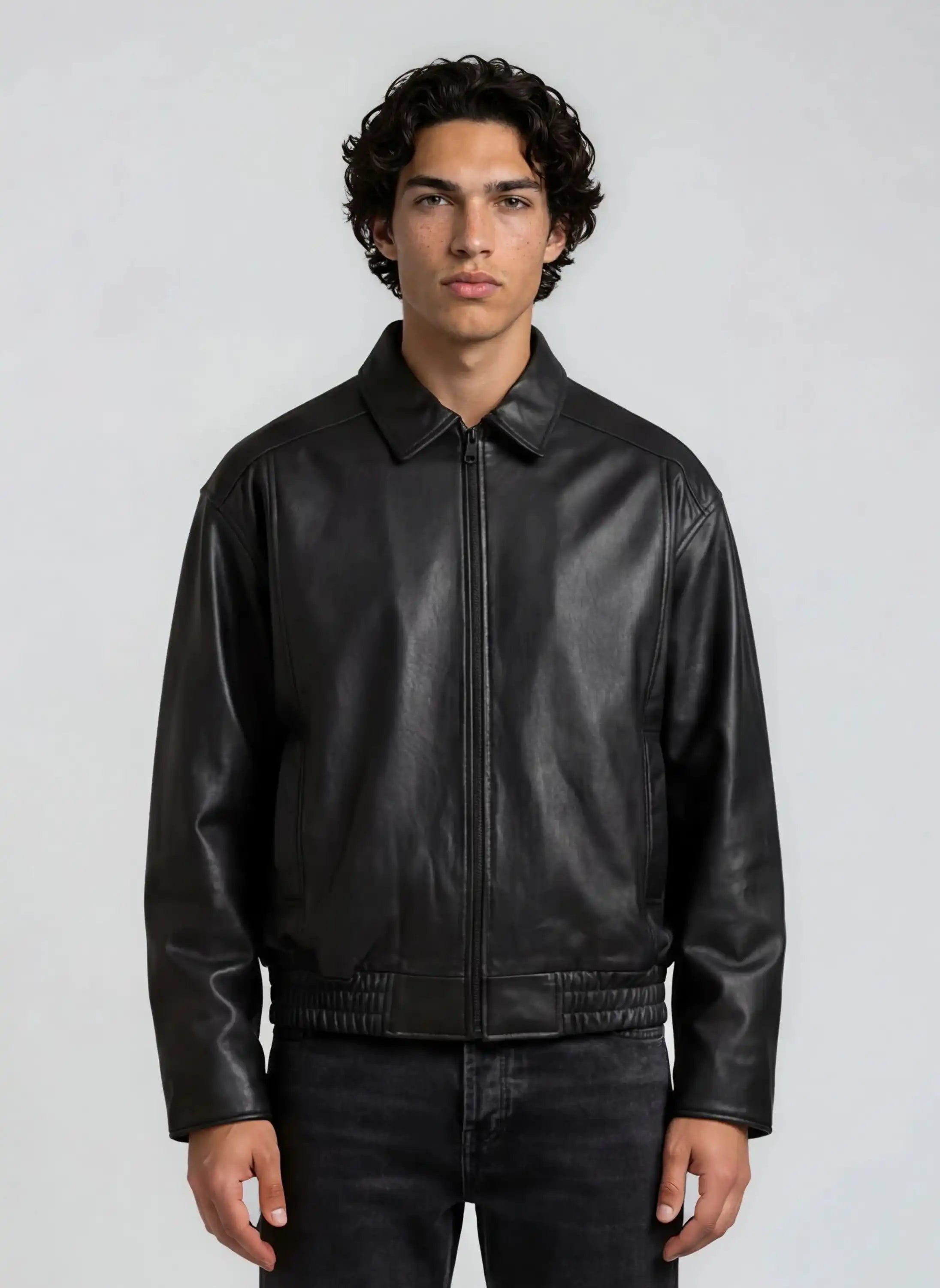 Eine Person mit kurzen lockigen Haaren trägt die trueprodigy Herren Lederjacke aus Schafsleder in Schwarz mit klassischem Blouson-Schnitt und schwarzen Jeans, steht vor einem schlichten hellgrauen Hintergrund und schaut in die Kamera.