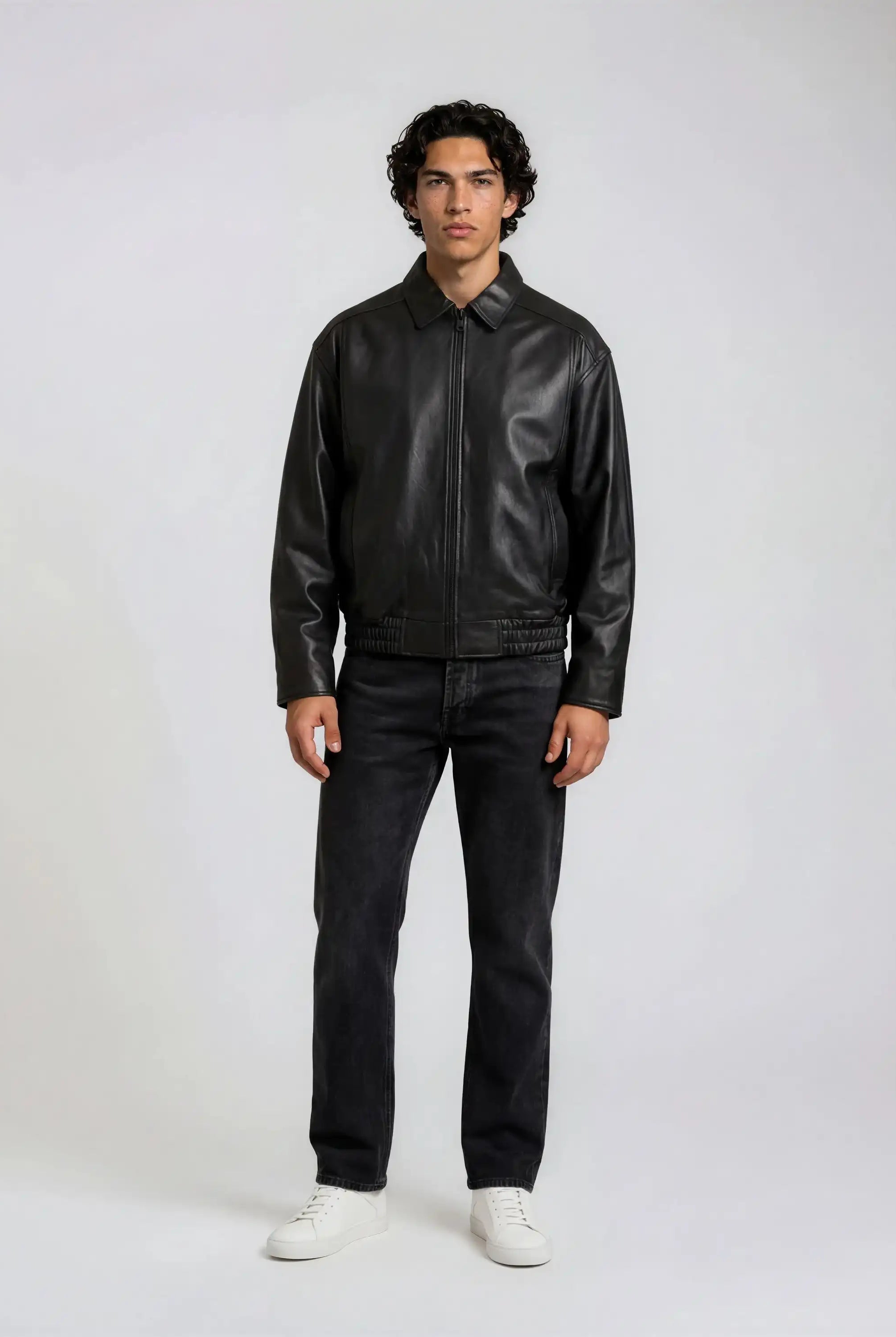 Ein Mann mit kurzem, lockigem Haar steht vor einem schlichten Hintergrund und trägt die trueprodigy Herren Lederjacke aus Schafsleder in Schwarz mit klassischem Blouson-Schnitt, dunkle Jeans und weiße Turnschuhe. Seine Arme liegen entspannt an den Seiten.