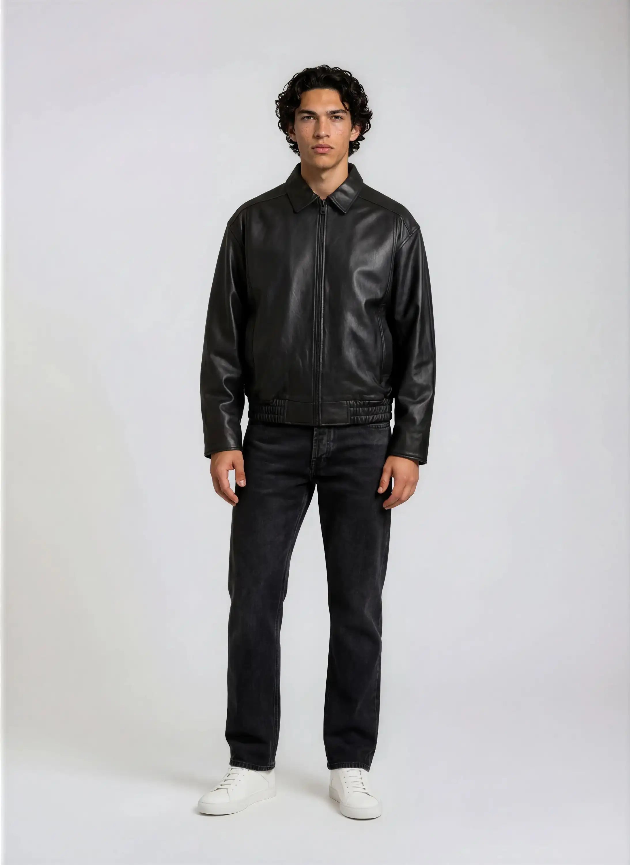 Ein Mann mit kurzem, lockigem Haar steht vor einem schlichten Hintergrund und trägt die trueprodigy Herren Lederjacke aus Schafsleder in Schwarz mit klassischem Blouson-Schnitt, dunkle Jeans und weiße Turnschuhe. Seine Arme liegen entspannt an den Seiten.