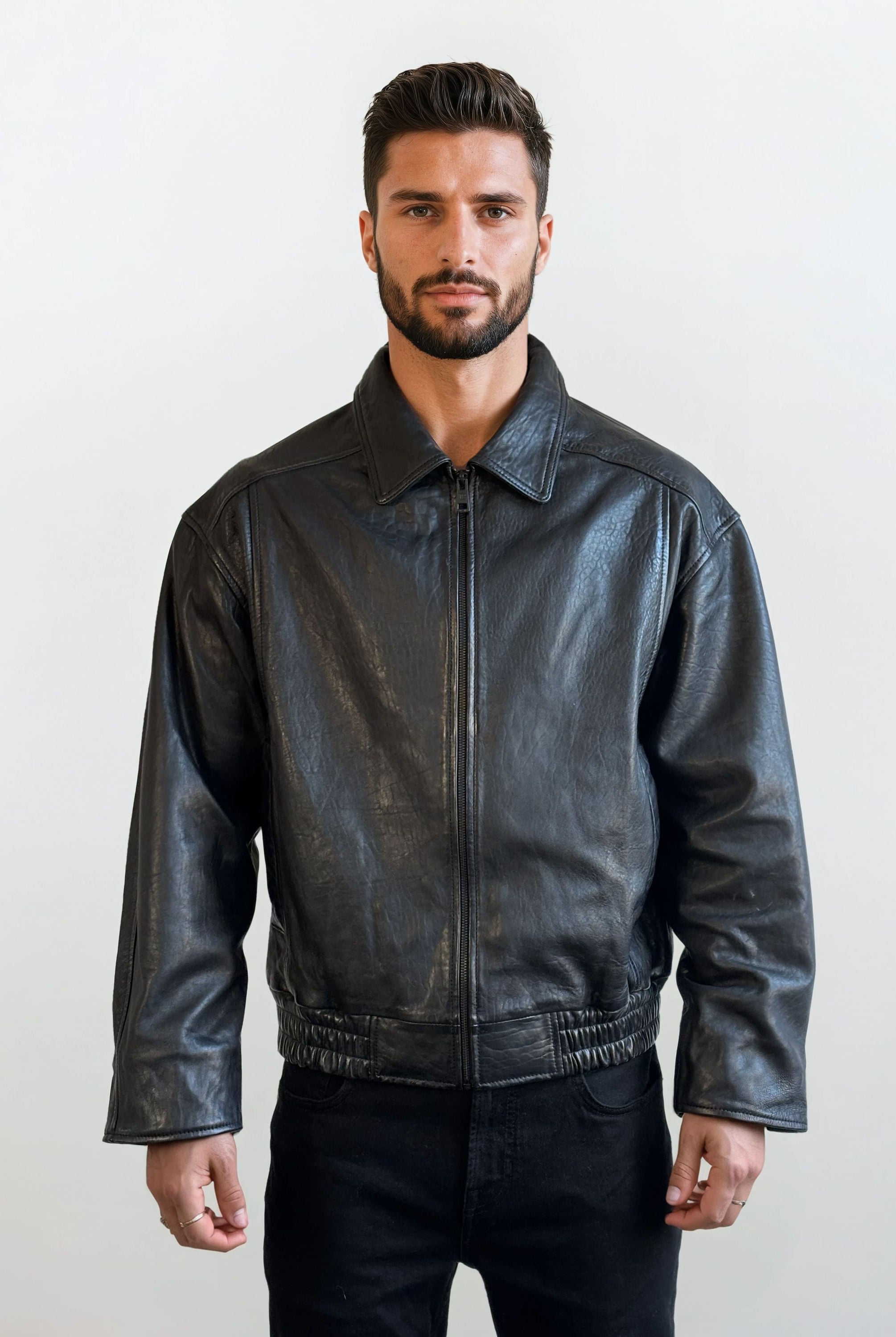 Ein Mann mit kurzen dunklen Haaren und einem Bart steht mit dem Gesicht nach vorne und trägt eine Trueprodigy Herren Lederjacke aus Schafsleder in Schwarz mit klassischem Blouson-Schnitt und eine schwarze Hose vor einem schlichten hellen Hintergrund.