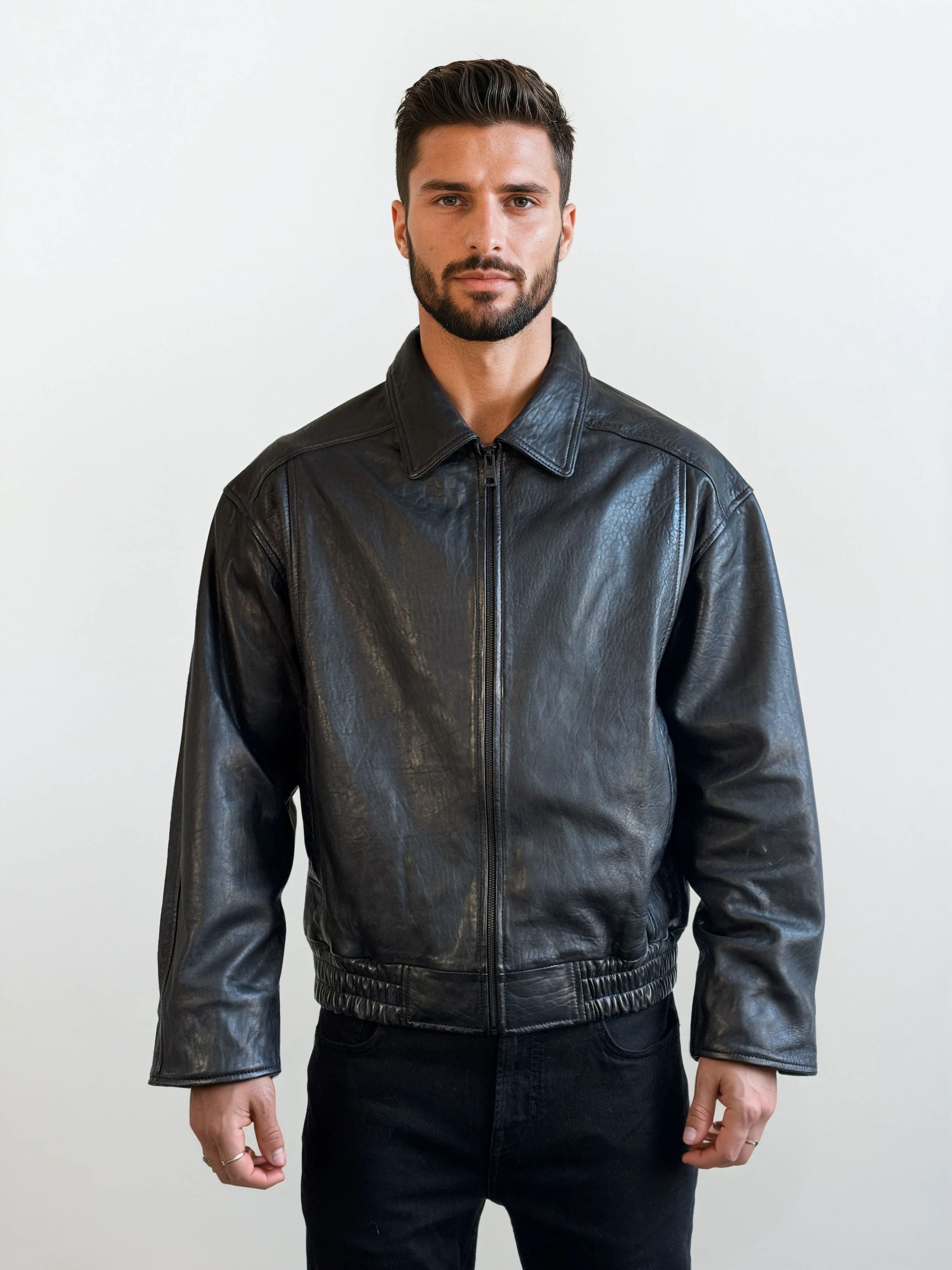 Ein Mann mit kurzen dunklen Haaren und einem Bart steht mit dem Gesicht nach vorne und trägt eine Trueprodigy Herren Lederjacke aus Schafsleder in Schwarz mit klassischem Blouson-Schnitt und eine schwarze Hose vor einem schlichten hellen Hintergrund.