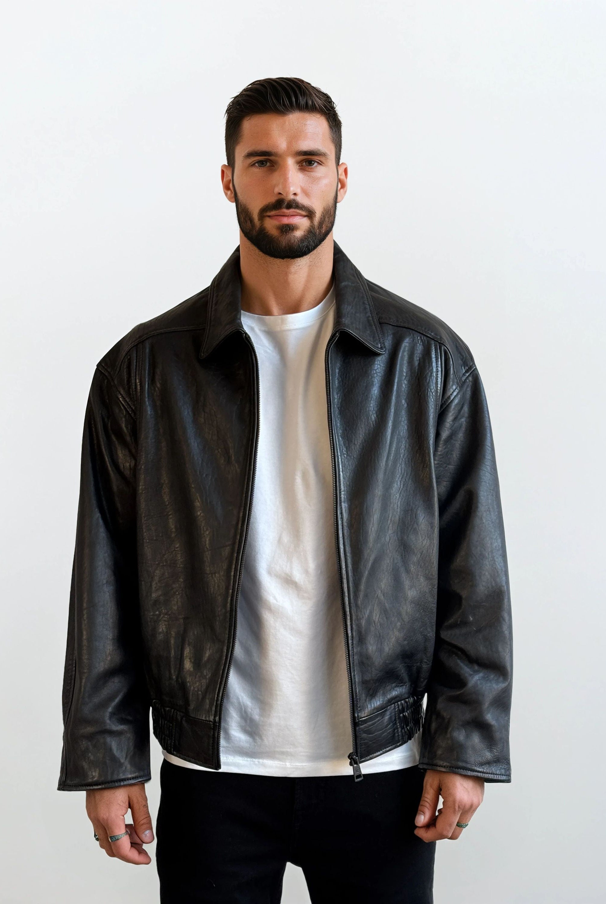 Ein Mann mit kurzen dunklen Haaren und einem Bart trägt die trueprodigy Herren Lederjacke aus Schafsleder in Schwarz mit klassischem Blouson-Schnitt über einem weißen T-Shirt und einer schwarzen Hose. Er steht vor einem schlichten weißen Hintergrund und schaut in die Kamera.