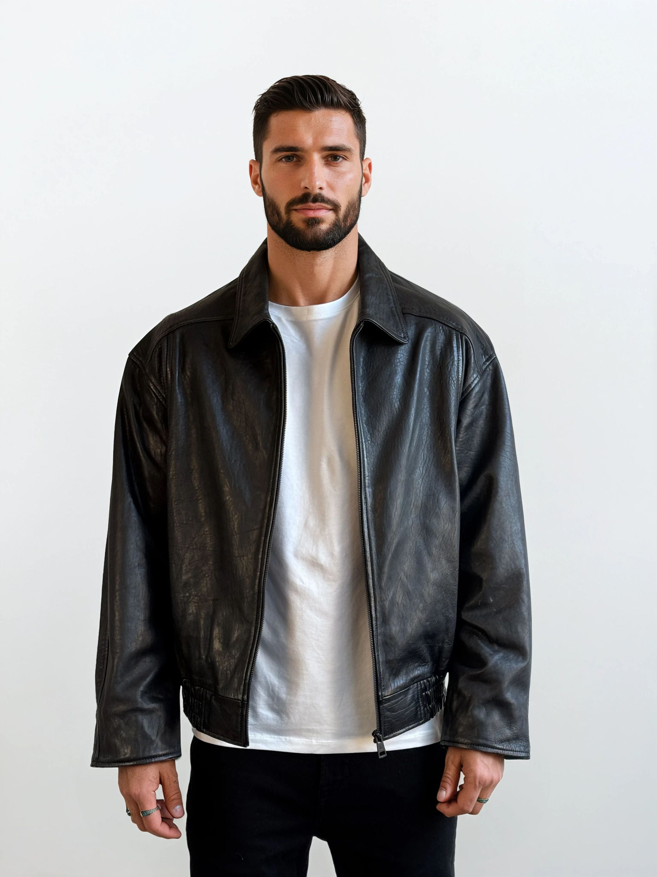 Ein Mann mit kurzen dunklen Haaren und einem Bart trägt die trueprodigy Herren Lederjacke aus Schafsleder in Schwarz mit klassischem Blouson-Schnitt über einem weißen T-Shirt und einer schwarzen Hose. Er steht vor einem schlichten weißen Hintergrund und schaut in die Kamera.