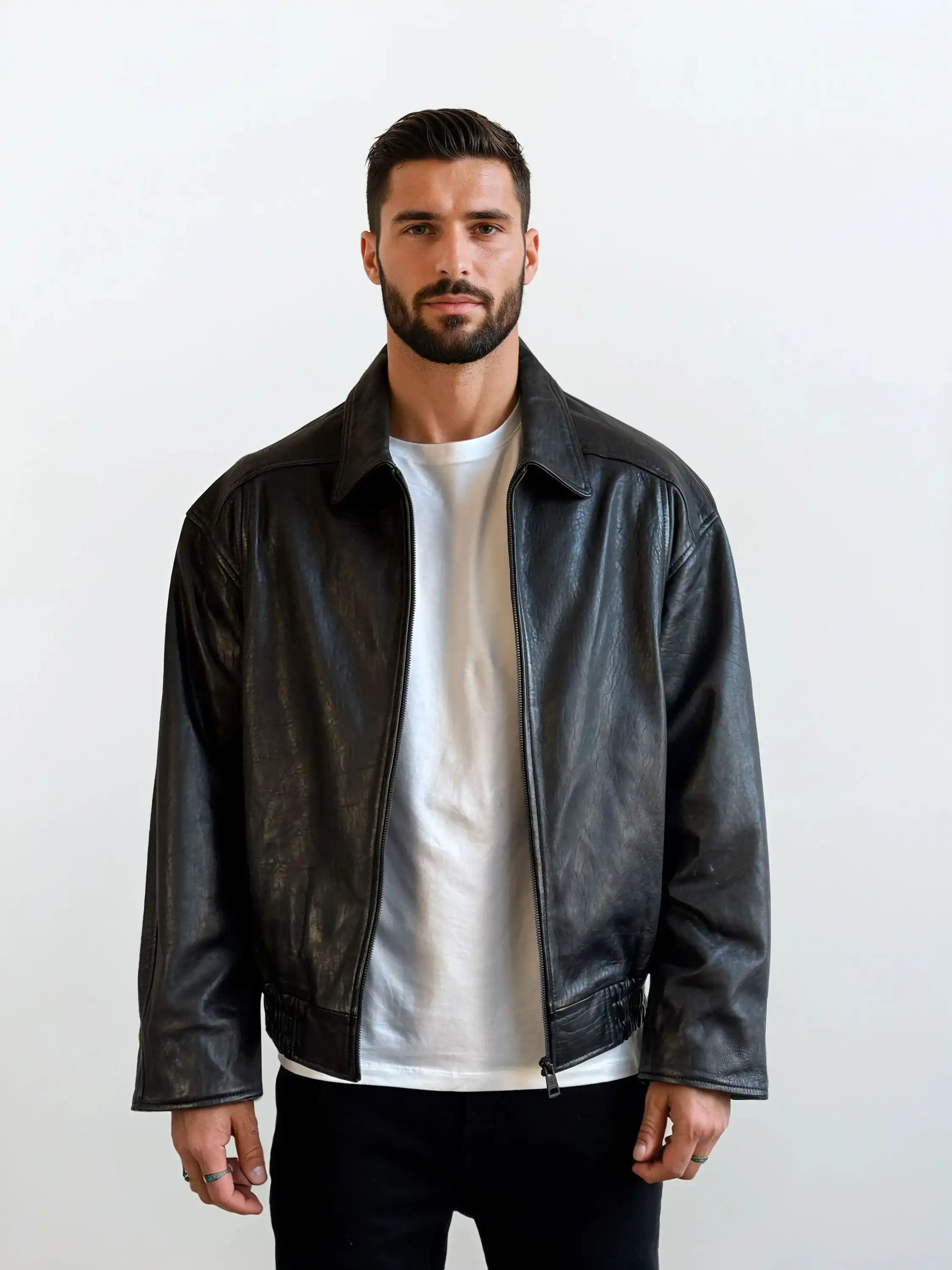 Ein Mann mit kurzen dunklen Haaren und einem Bart trägt die trueprodigy Herren Lederjacke aus Schafsleder in Schwarz mit klassischem Blouson-Schnitt über einem weißen T-Shirt und einer schwarzen Hose. Er steht vor einem schlichten weißen Hintergrund und schaut in die Kamera.