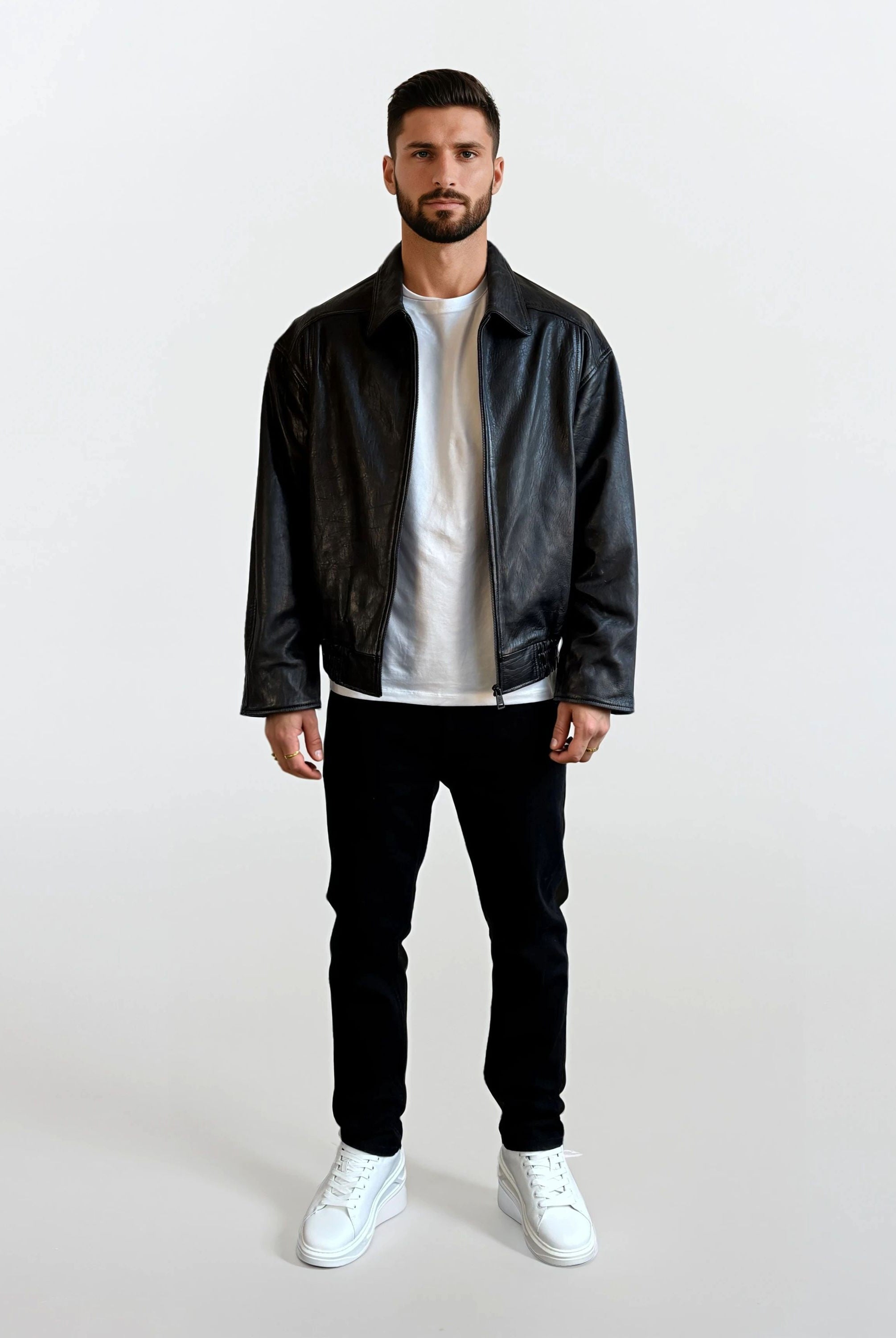 Ein Mann mit kurzen dunklen Haaren und einem Bart, der eine trueprodigy Herren Lederjacke aus Schafsleder in Schwarz mit klassischem Blouson-Schnitt, ein weißes T-Shirt, eine schwarze Hose und weiße Turnschuhe trägt, steht vor einem schlichten weißen Hintergrund.