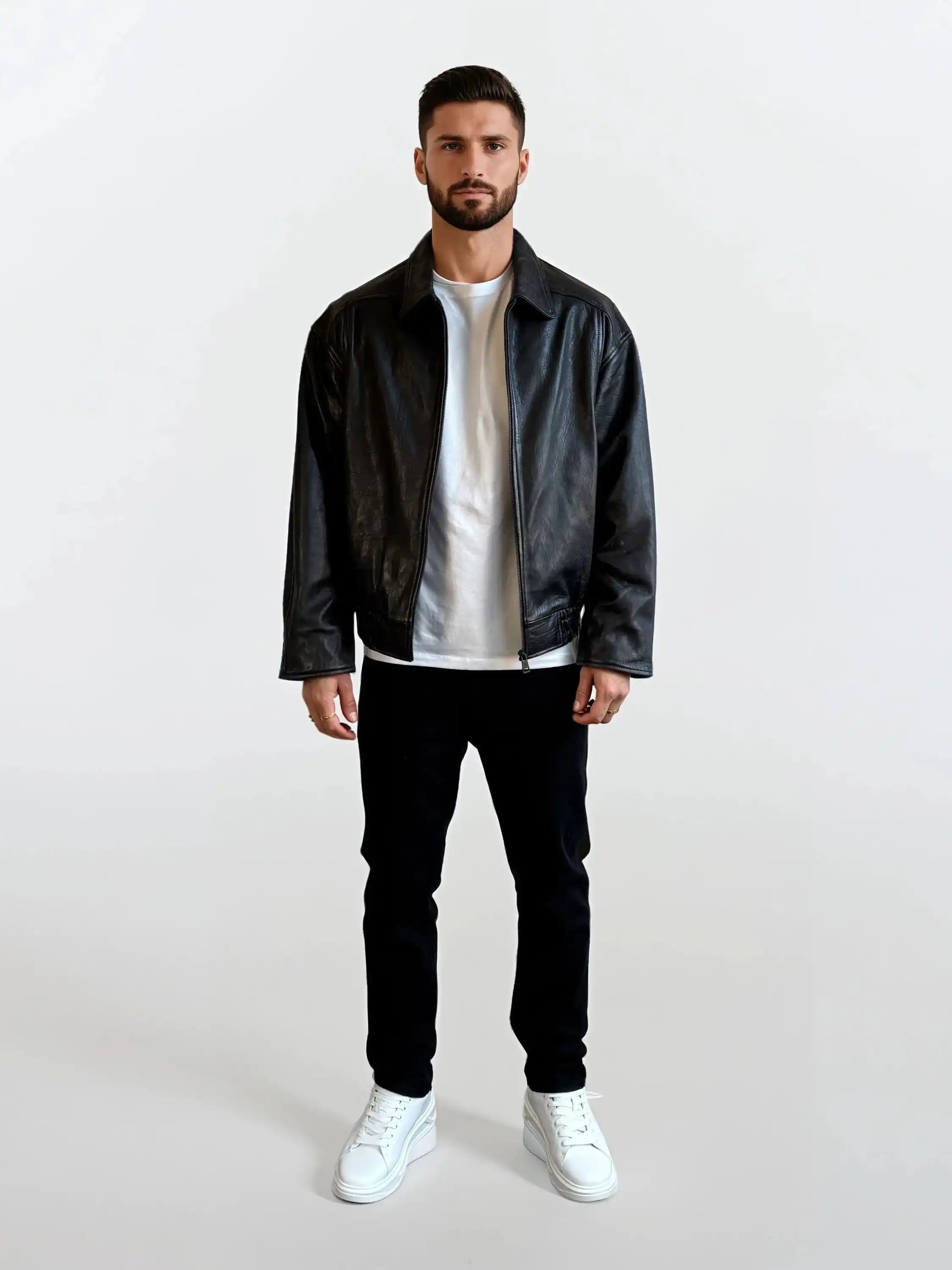 Ein Mann mit kurzen dunklen Haaren und einem Bart, der eine trueprodigy Herren Lederjacke aus Schafsleder in Schwarz mit klassischem Blouson-Schnitt, ein weißes T-Shirt, eine schwarze Hose und weiße Turnschuhe trägt, steht vor einem schlichten weißen Hintergrund.