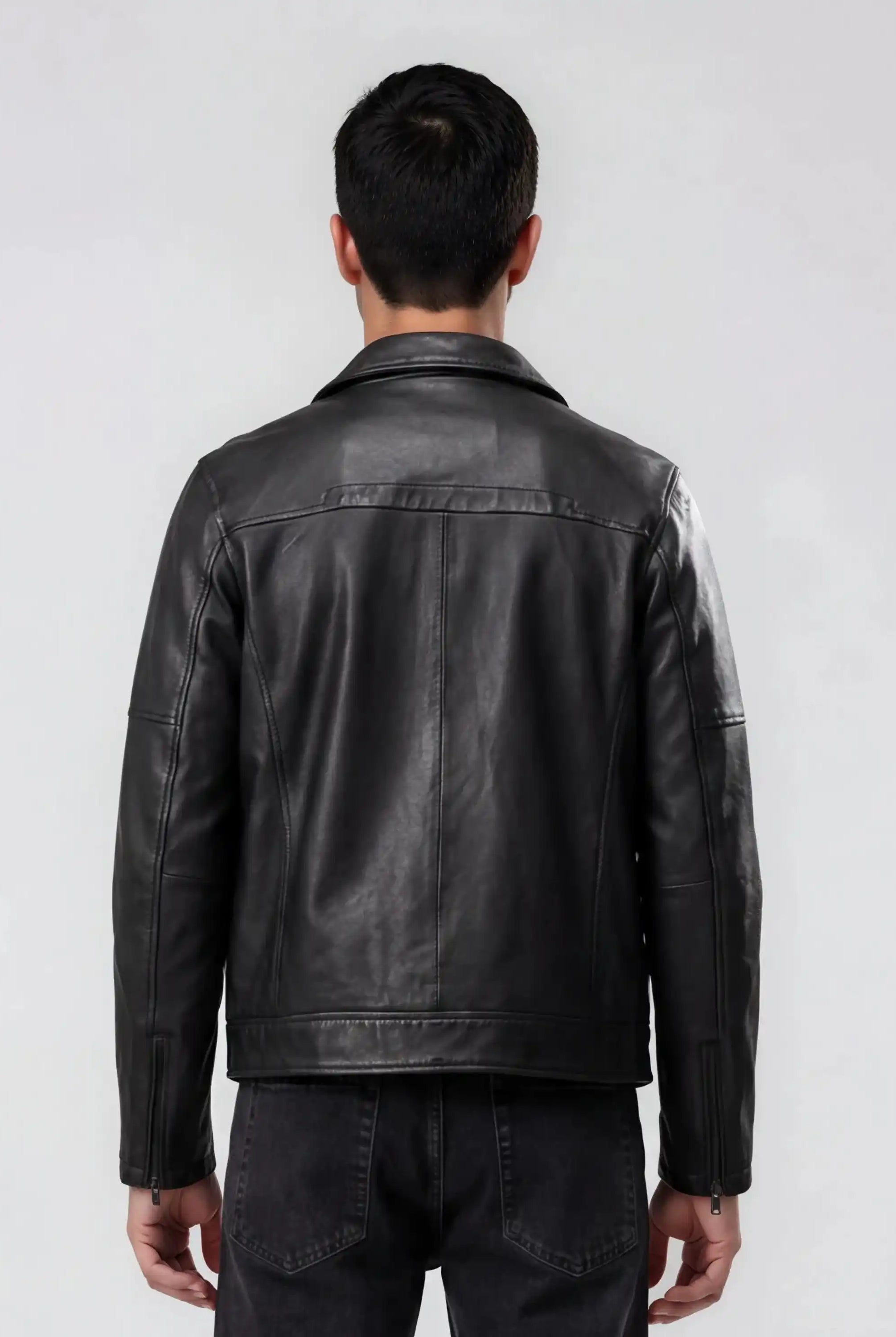 Ein Mann steht mit dem Rücken zur Kamera und trägt eine trueprodigy Herren Lederjacke aus Schafsleder in Schwarz mit Ärmel-Reißverschlüssen und dunkle Jeans vor einem schlichten hellgrauen Hintergrund.