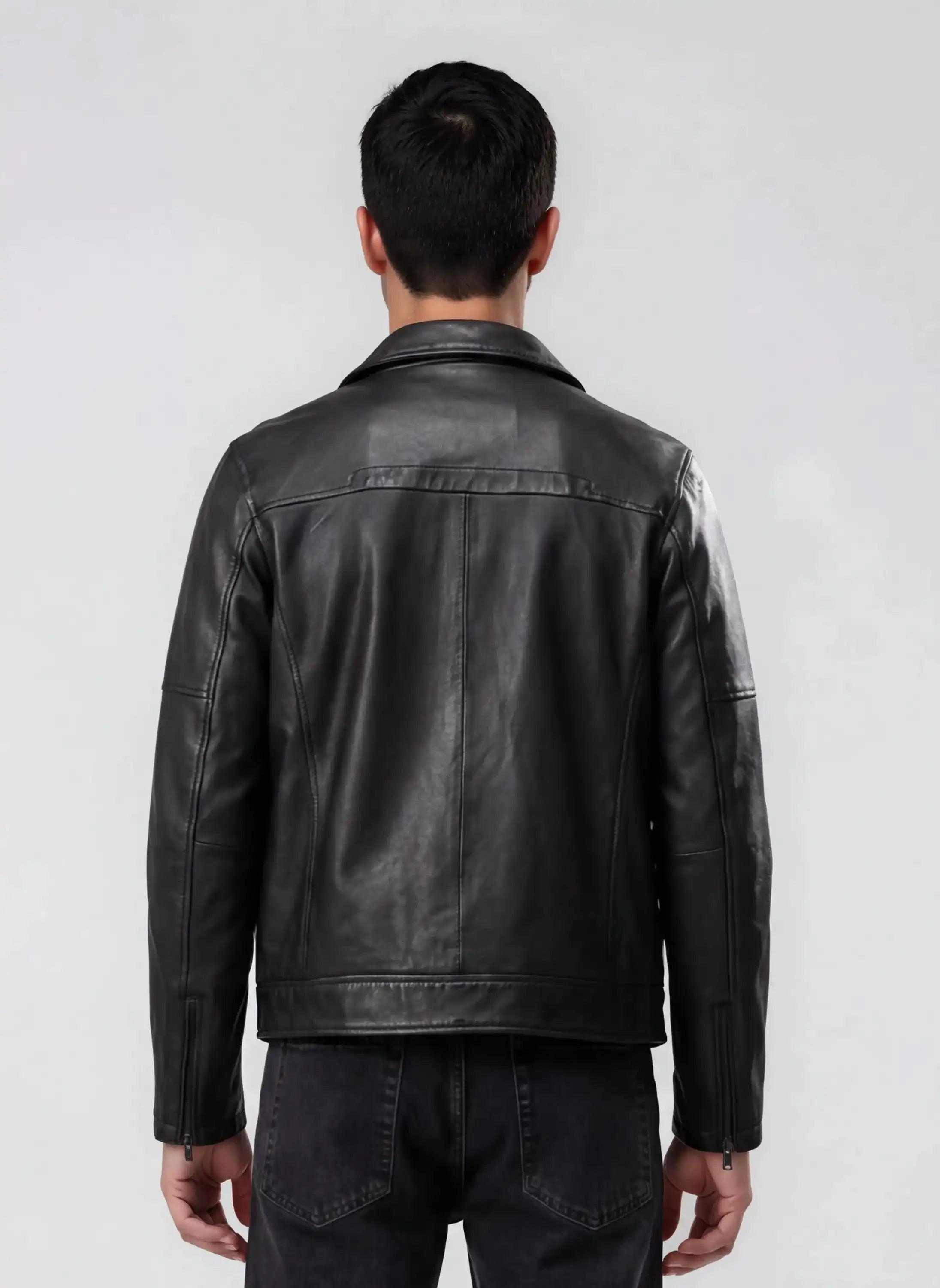 Ein Mann steht mit dem Rücken zur Kamera und trägt eine trueprodigy Herren Lederjacke aus Schafsleder in Schwarz mit Ärmel-Reißverschlüssen und dunkle Jeans vor einem schlichten hellgrauen Hintergrund.