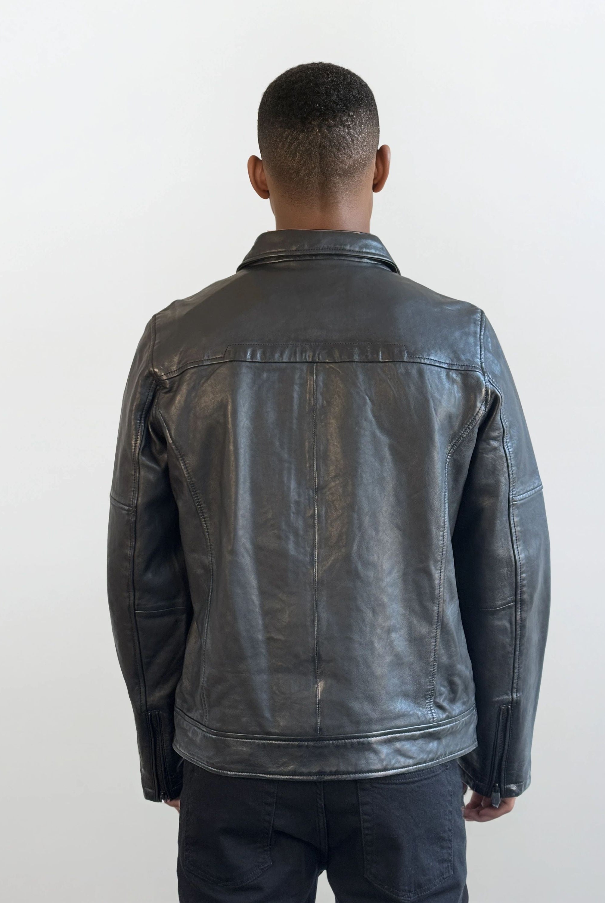 Man sieht eine Person mit kurzen Haaren von hinten, die eine trueprodigy Herren Lederjacke aus Schafsleder in Schwarz mit Ärmel-Reißverschlüssen und eine dunkle Hose trägt und vor einem schlichten, hellen Hintergrund steht.