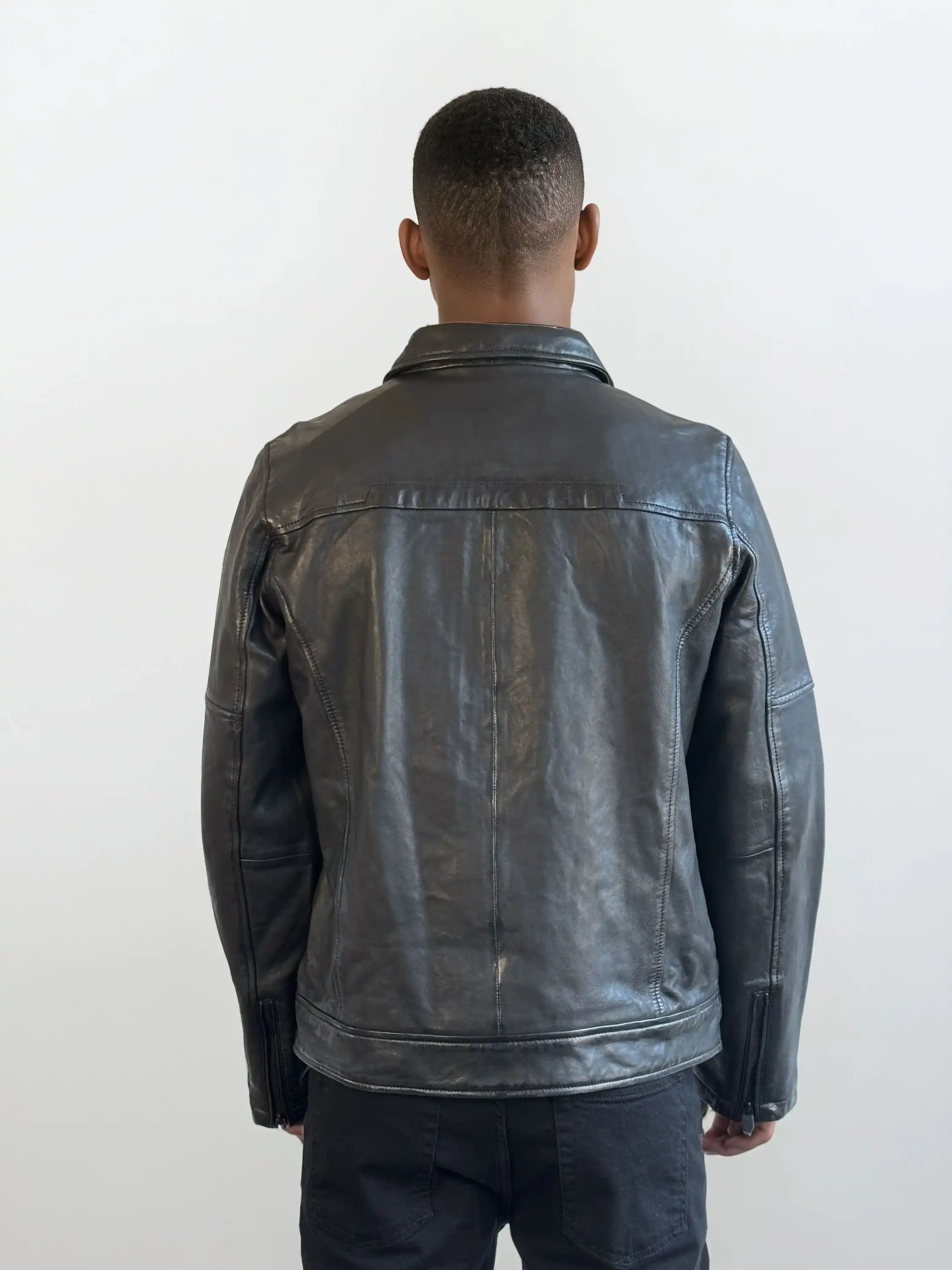 Man sieht eine Person mit kurzen Haaren von hinten, die eine trueprodigy Herren Lederjacke aus Schafsleder in Schwarz mit Ärmel-Reißverschlüssen und eine dunkle Hose trägt und vor einem schlichten, hellen Hintergrund steht.