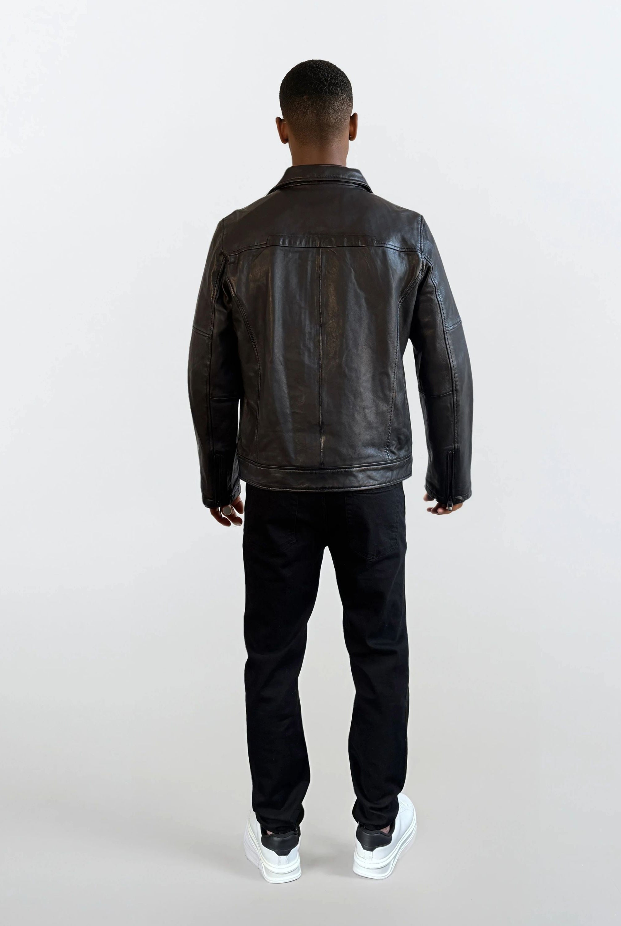 Eine Person trägt eine schwarze Herren Lederjacke aus Schafsleder mit Ärmel-Reißverschlüssen von trueprodigy, dazu schwarze Hosen und weiße Sneaker vor einem schlichten, hellen Hintergrund.